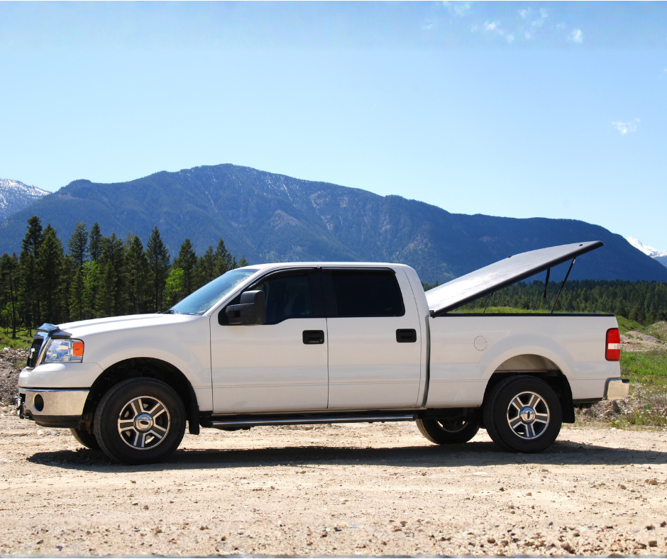 White pickup truck with a raised tailgate set against mountainous terrain and forested landscape under a clear blue sky.