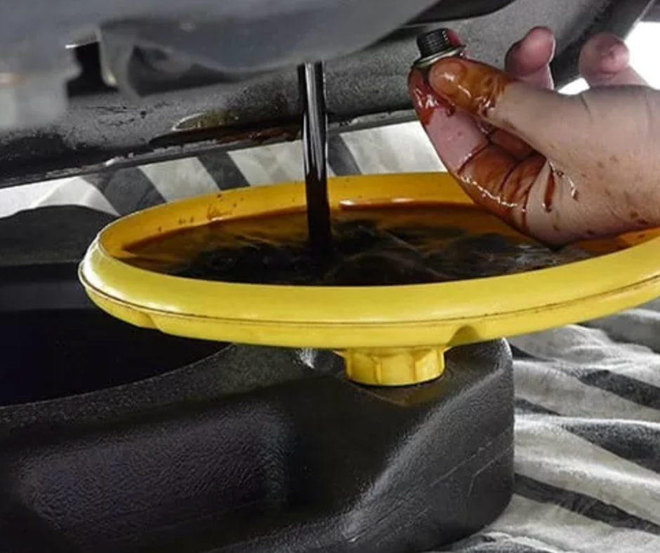 A person pouring dark engine oil into a yellow oil catch pan underneath a vehicle's oil pan, with their hand covered in oil.