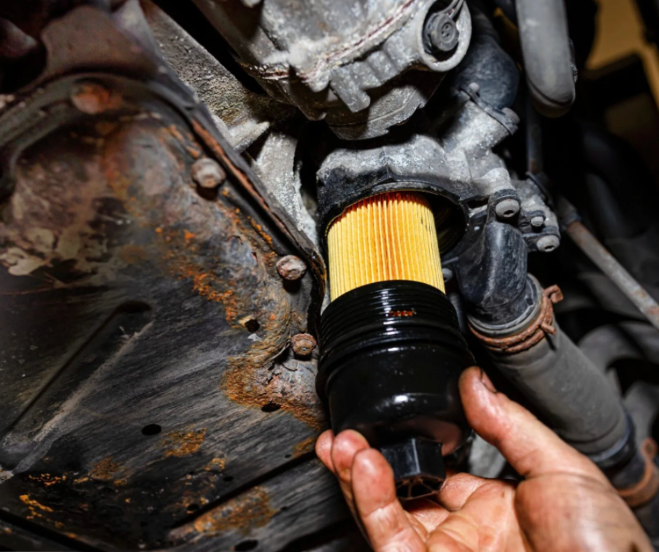 Close-up of a person replacing the oil filter on a vehicle engine, with visible rust and mechanical parts in the background.