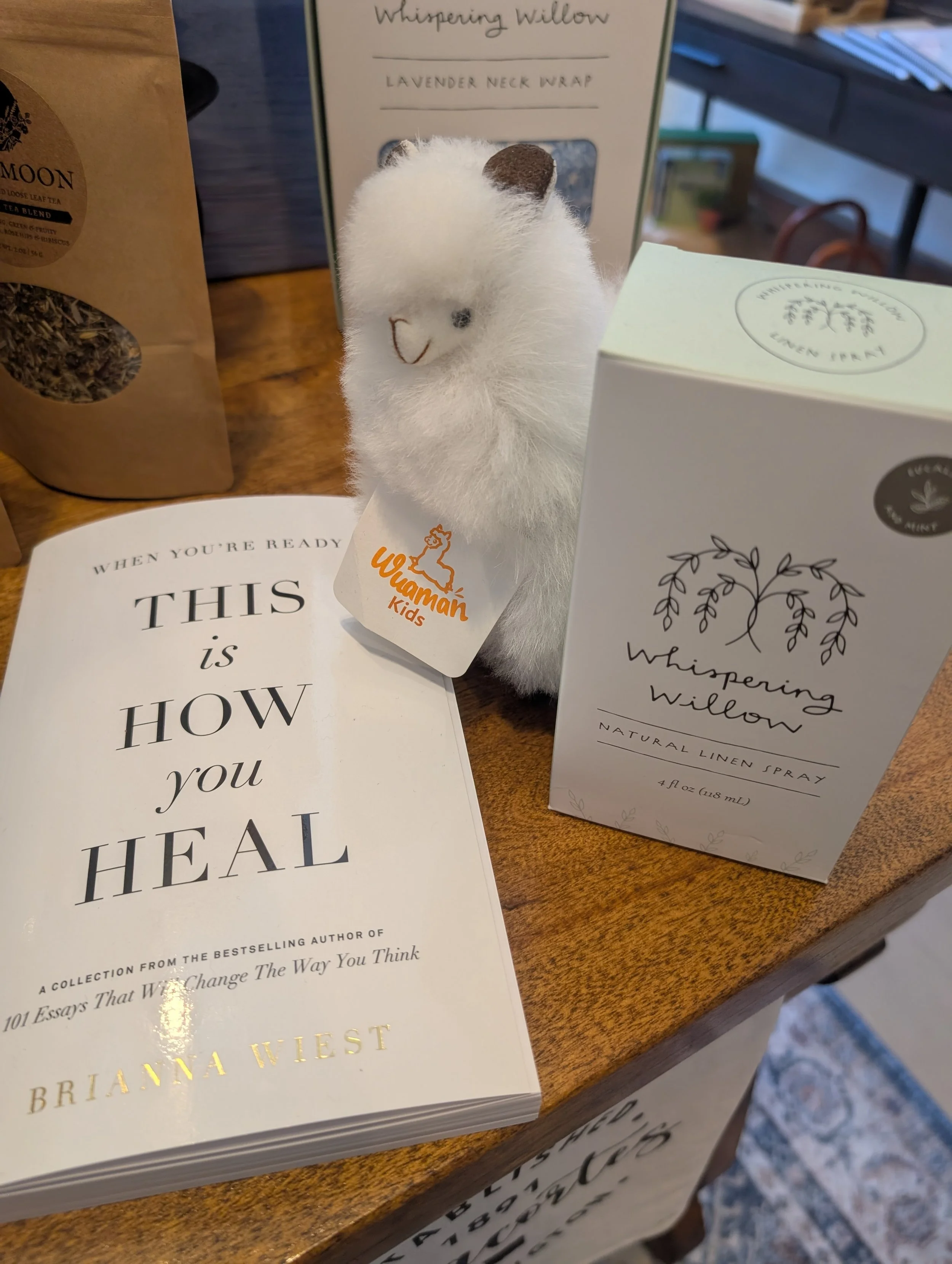 A plush white llama stuffed animal with a tag that says 'Wamama Kids' on a wooden table. Next to it a book titled 'This Is How You Heal' by Brianna Wiest. Behind the llama, there's a box labeled 'Whispering Willow Lavender Neck Wrap' and a box labeled 'Whispering Willow Natural Linen Spray.'