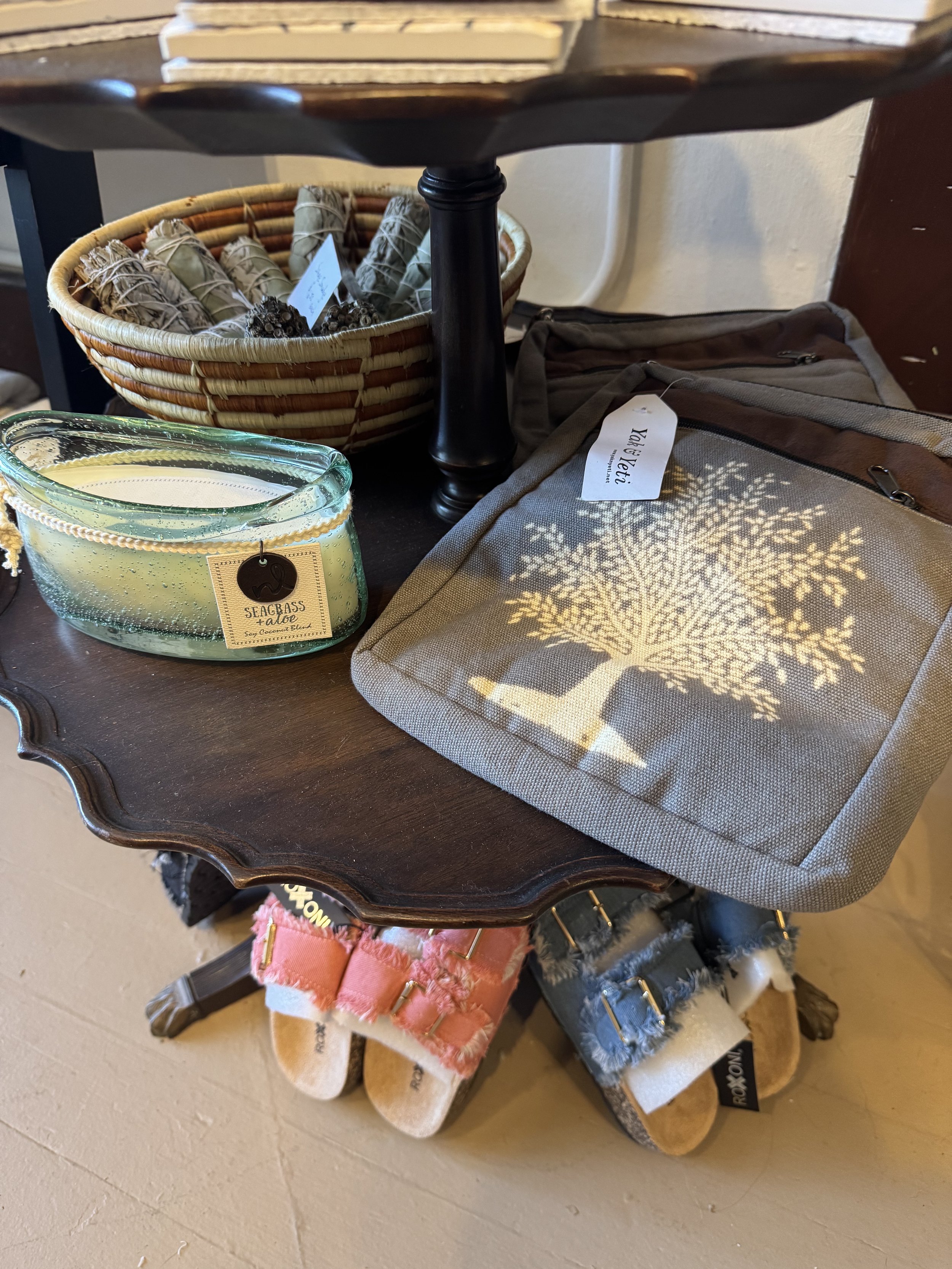 A dark wooden table displaying a wicker basket with lavender bundles, a glass dish candle labeled 'Seabass Talao,' a gray tote bag with a tree shadow pattern, a set of slippers with pink and gray frayed straps, and a small dark table with a black pedestal base.
