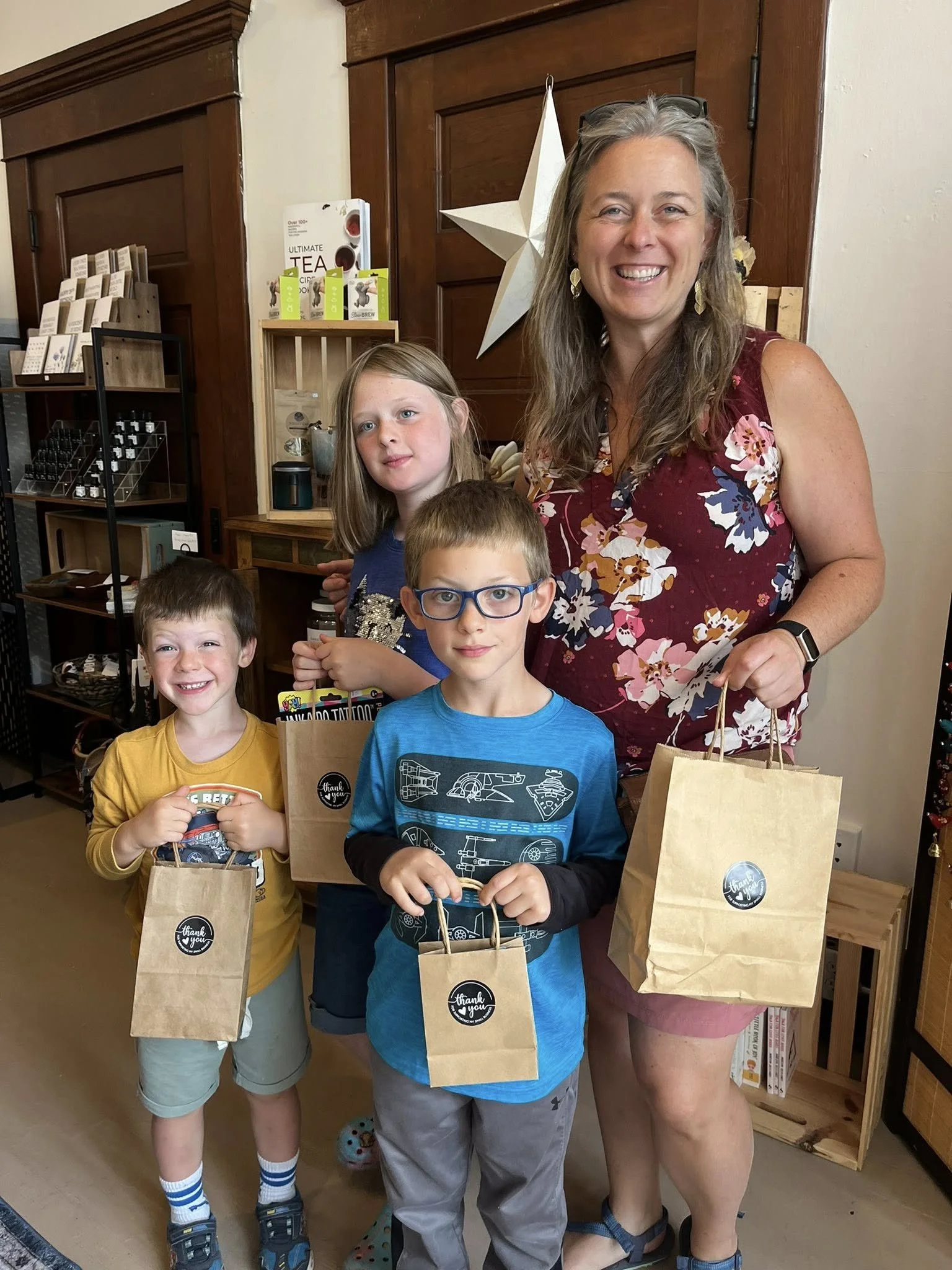 A smiling woman with three children holding small thank you gift bags standing inside a store with wood paneling and shelves of products.