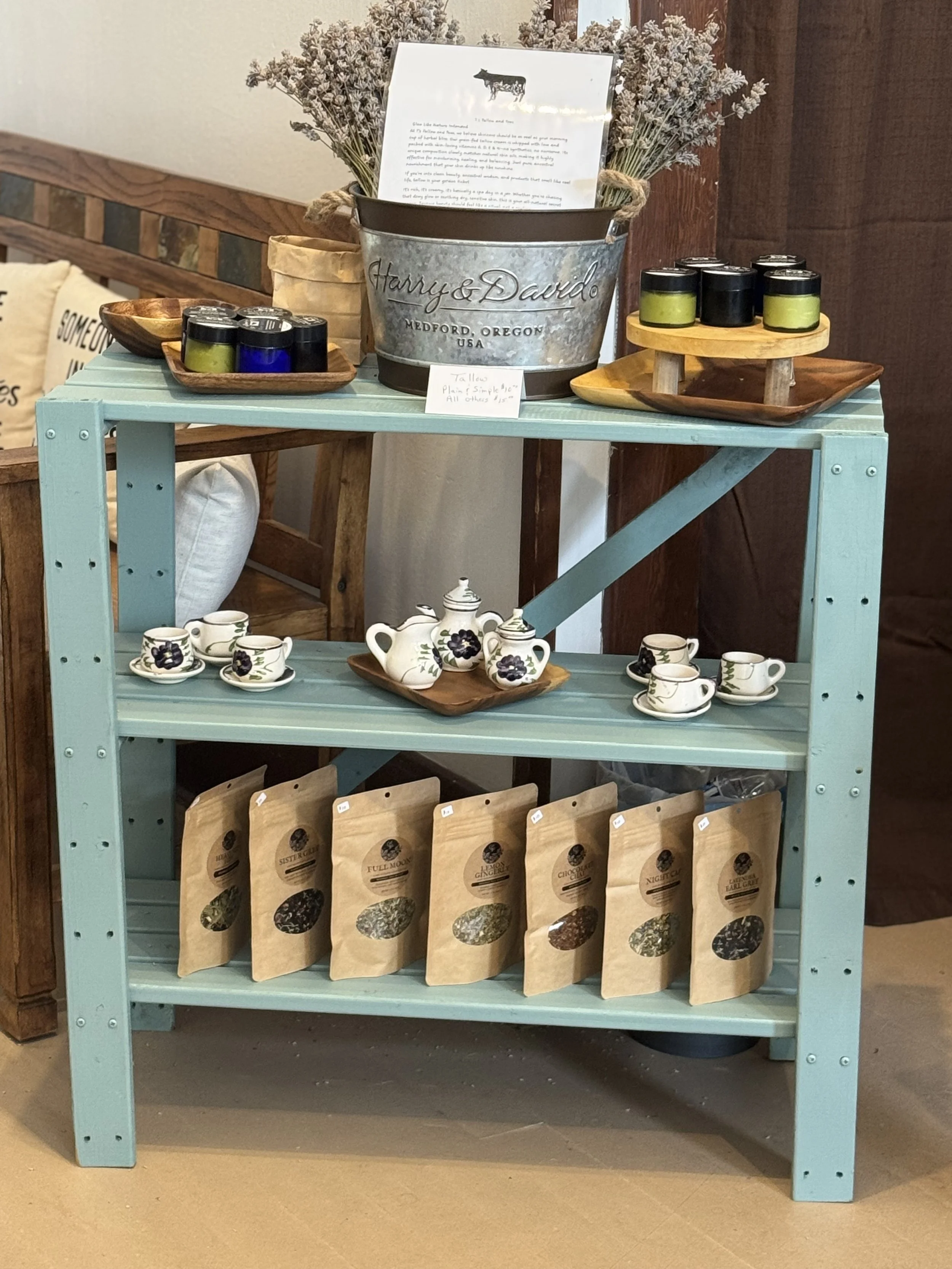 A blue wooden shelf displays a metal pail with dried flowers and a note, small trays with candles, a wooden tray with teacups and teapots decorated with purple flowers, and paper bags of loose tea on the bottom shelf.