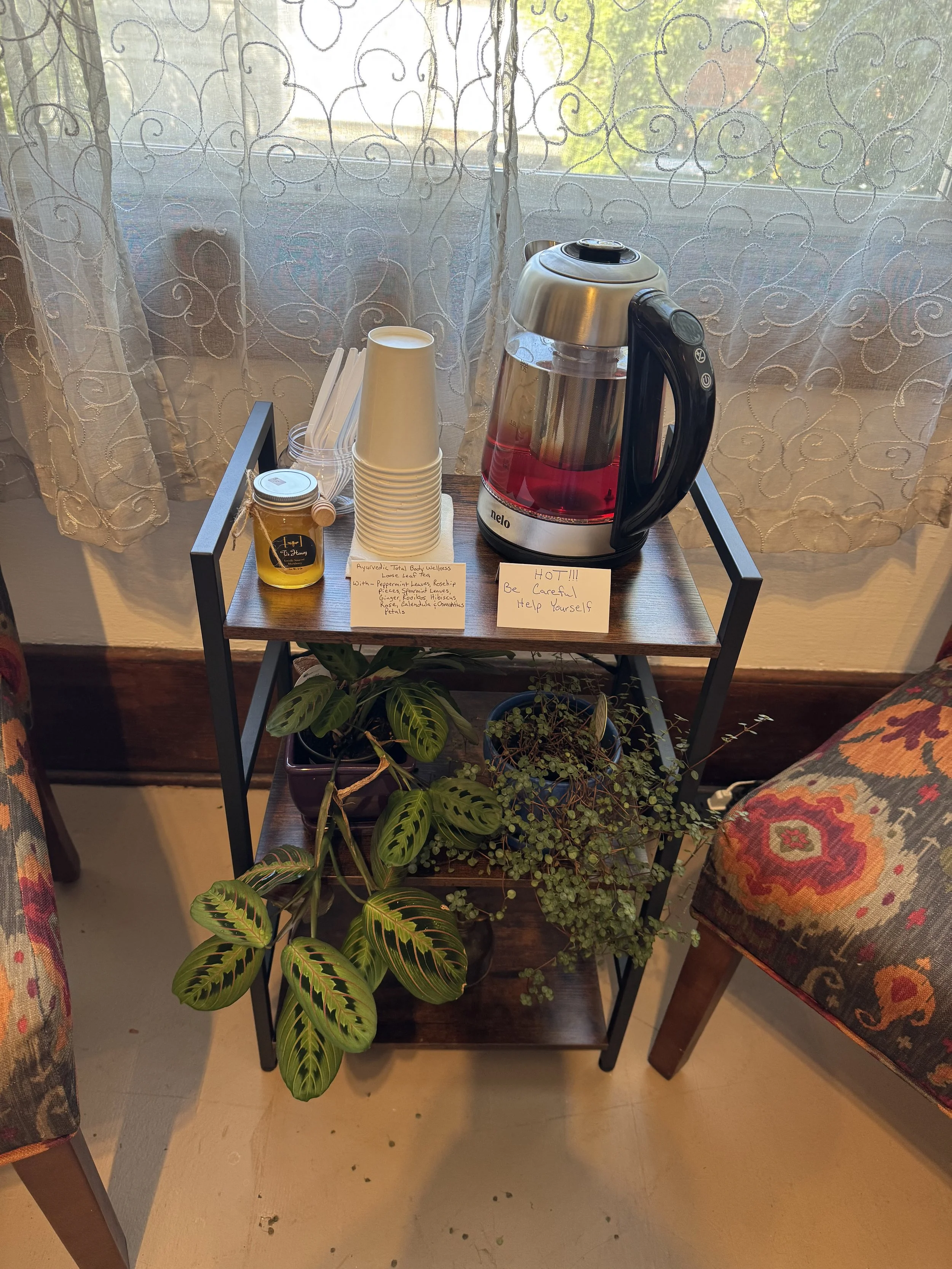 A small wooden table with a glass pitcher filled with red liquid, disposable cups, honey jar, and a note. Two patterned chairs are on each side, and potted plants are underneath the table. Behind the table is a window with lace curtains.