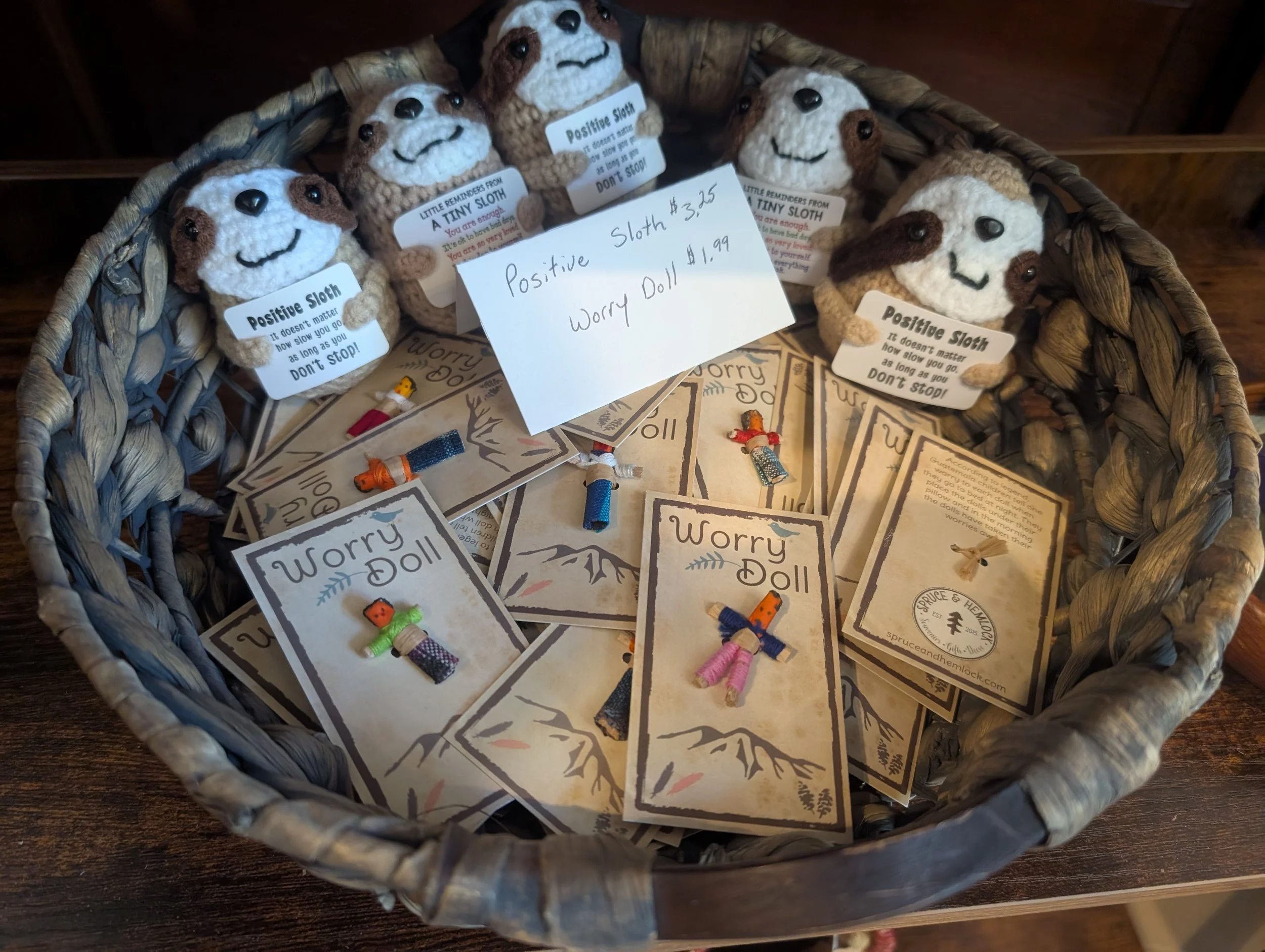 A basket filled with plush sloth toys labeled 'Positive Sloth' and handmade worry dolls on cards. The sloth toys have smiling faces, and the worry dolls are small with colorful string bodies. A handwritten sign indicates the sloth toy costs $3.25 and the worry doll costs $1.99.