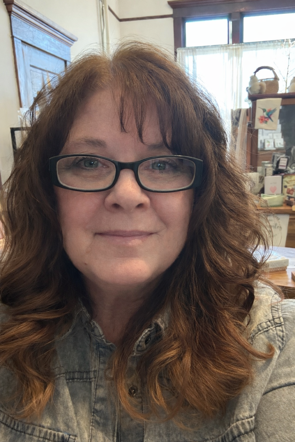 A woman with wavy, shoulder-length auburn hair wearing black glasses and a light denim jacket, smiling indoors with a background showing a window, curtains, and shelves with decorative items.