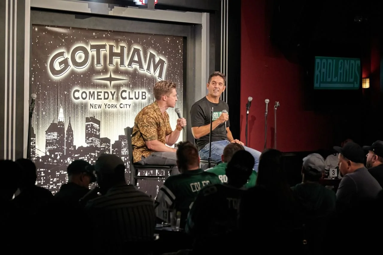 Two comedians perform on stage at Gotham Comedy Club in New York City, with a backdrop displaying the club's name and city. Audience members sit in front, watching the show.