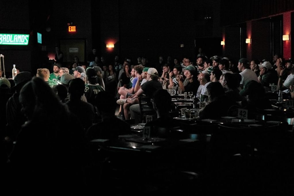 Audience watching a performance on stage in a dark comedy club with dim lighting and green exit sign.