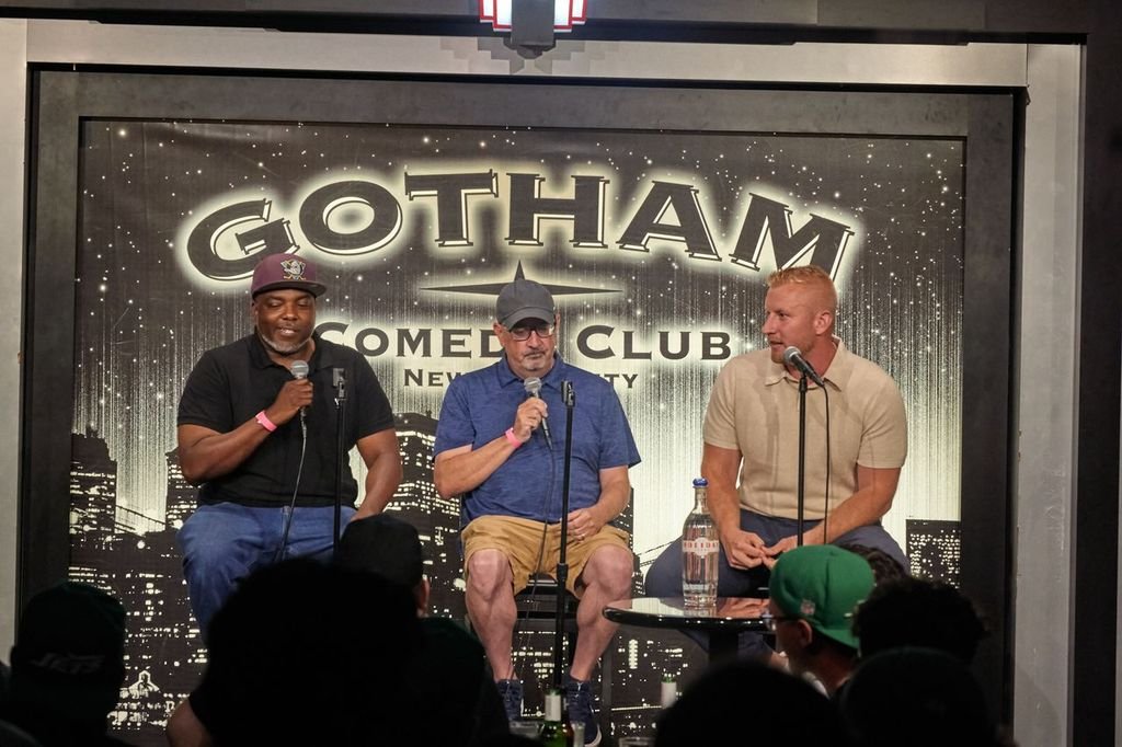 Three men sitting on stage at Gotham Comedy Club in New York City, holding microphones, with an audience visible in the foreground. The background features a cityscape and the Gotham Comedy Club logo.