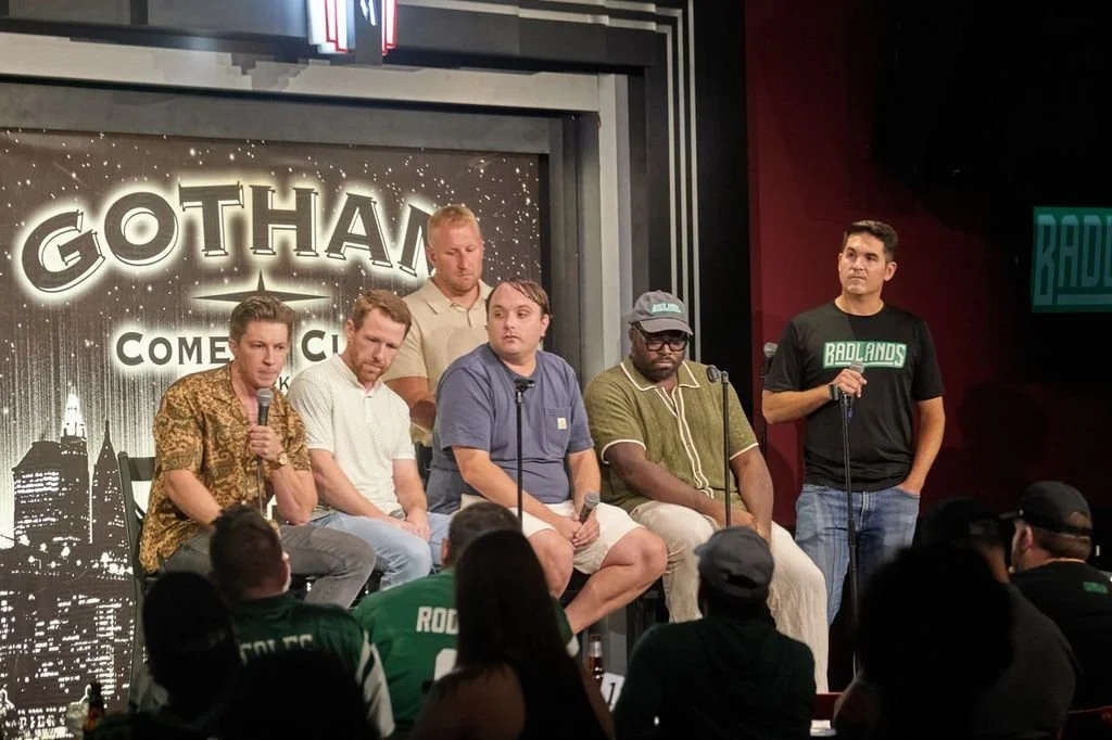 Six men are on a stage at Gotham Comedy Club, with four sitting and two standing, holding microphones, during a stand-up comedy event or panel discussion. The audience is visible in front.