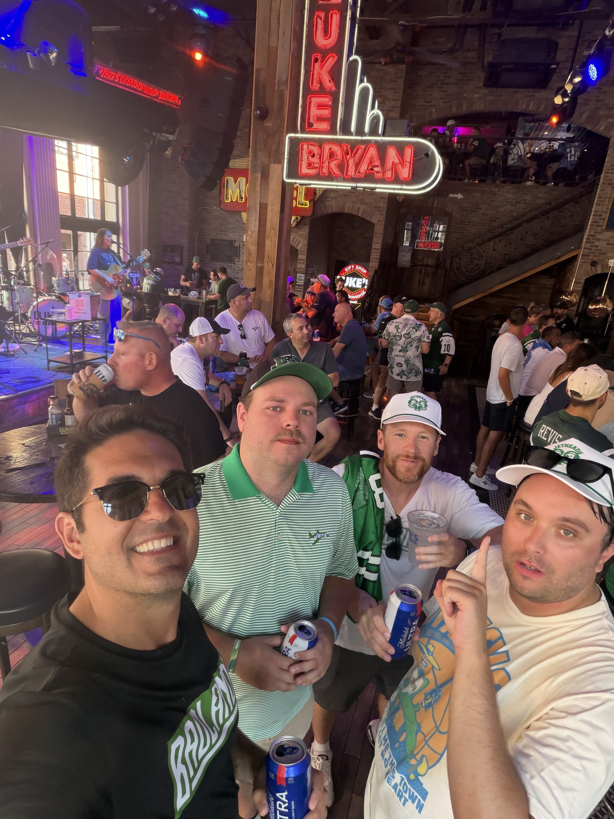 A group of people at a bar or pub during a sports game, some holding drinks, with a musician playing guitar and drums on stage in the background, neon signs, and brick walls.