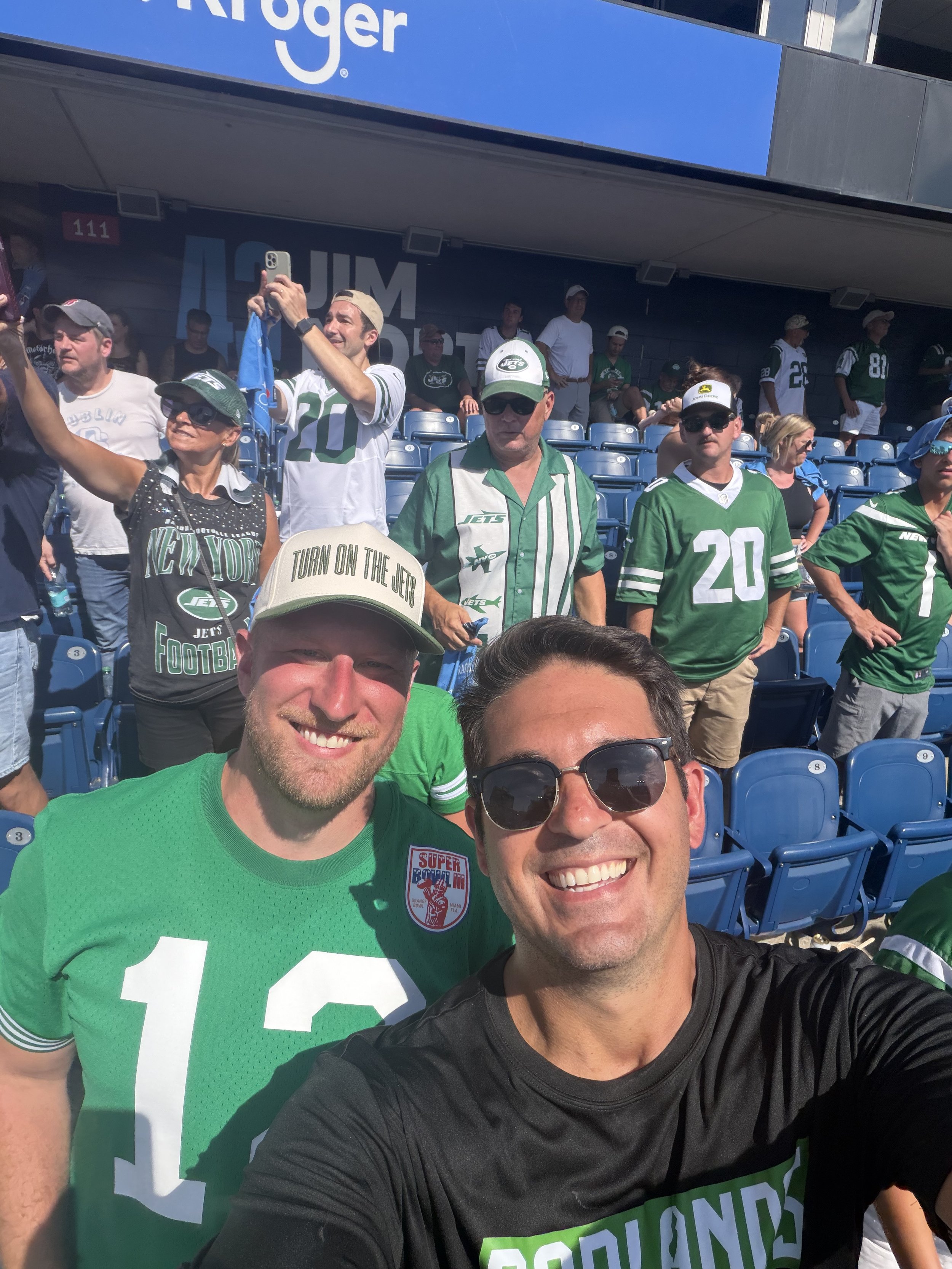 A group of sports fans at a stadium, many wearing New York Jets jerseys and hats, taking a selfie and enjoying a game.