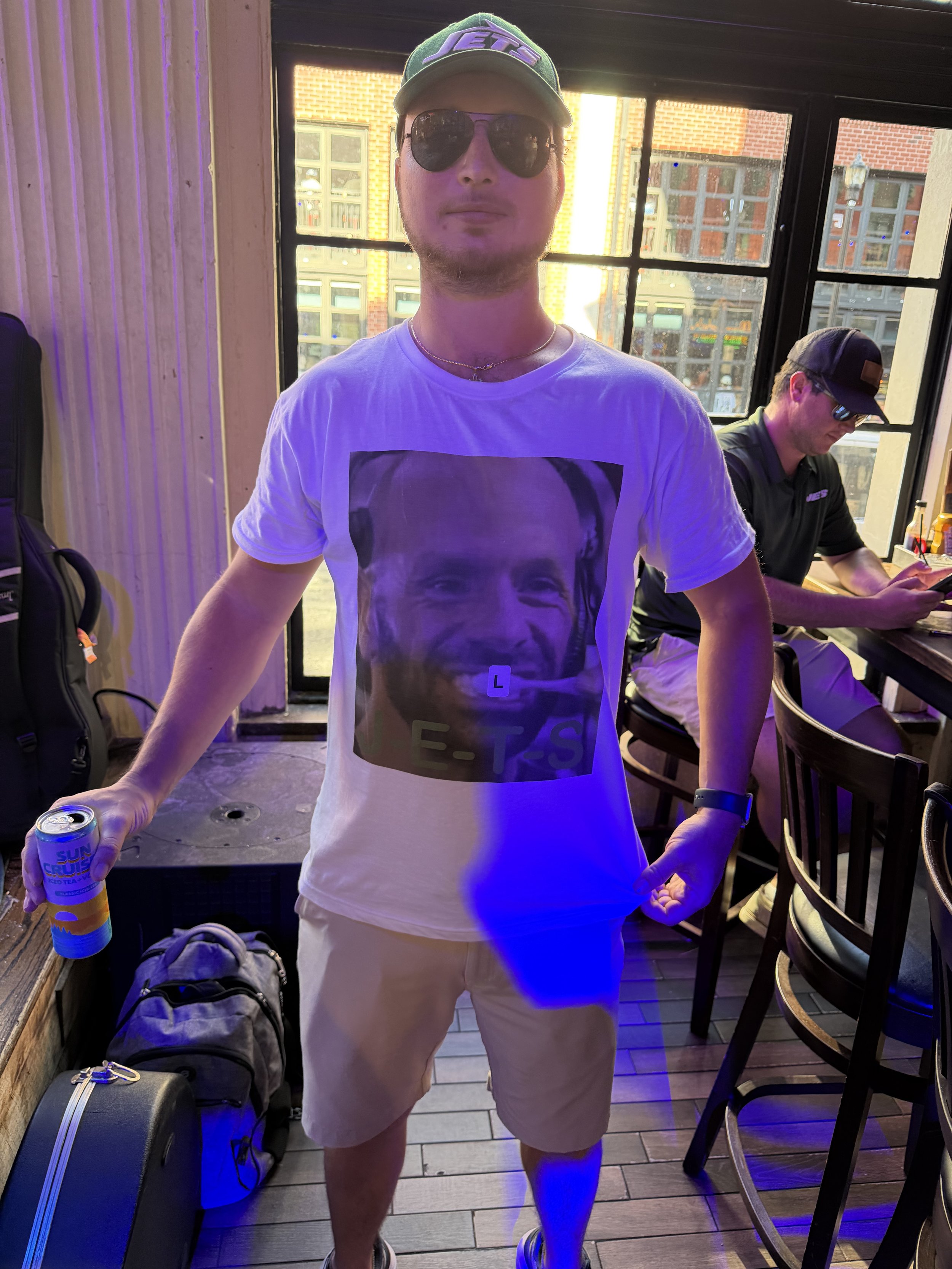 A man wearing sunglasses, a cap, a white graphic t-shirt, khaki shorts, and sneakers stands in a cafe or restaurant, holding a can of Sun Crush iced tea. He has a backpack and a suitcase at his side. A person dressed in black with sunglasses and a ca