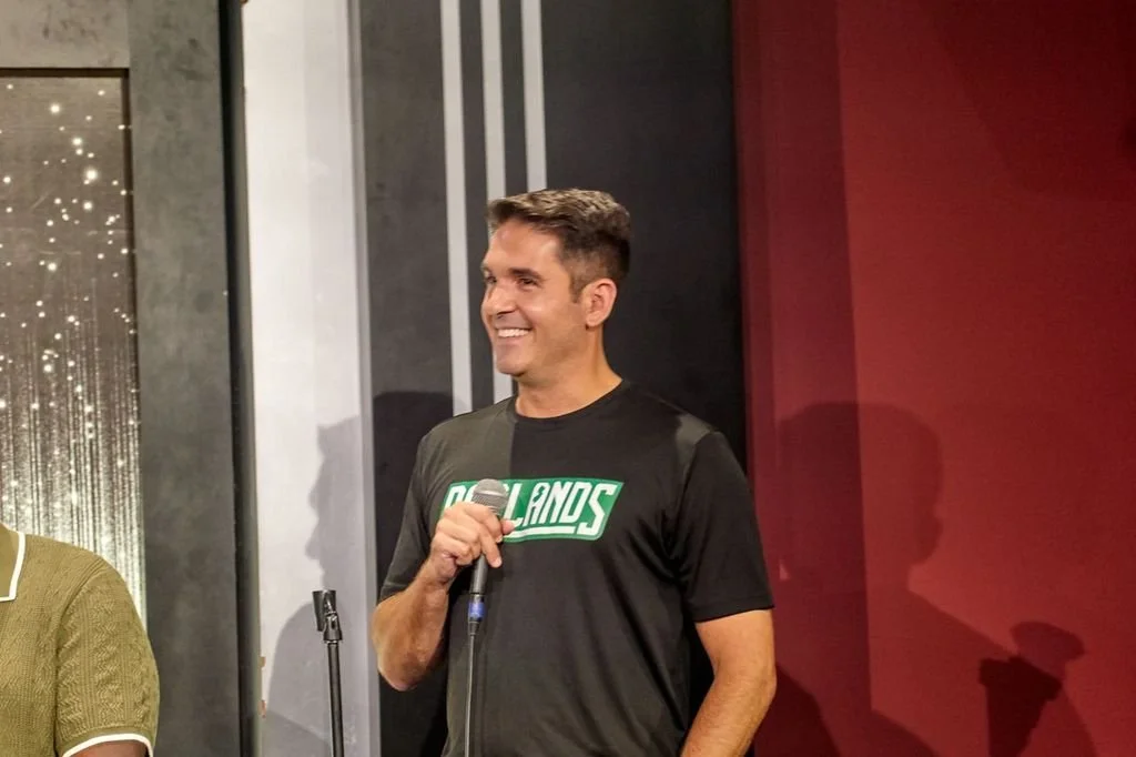 A man smiling and holding a microphone, wearing a black t-shirt with the words 'Orlando Magic' in green and white.