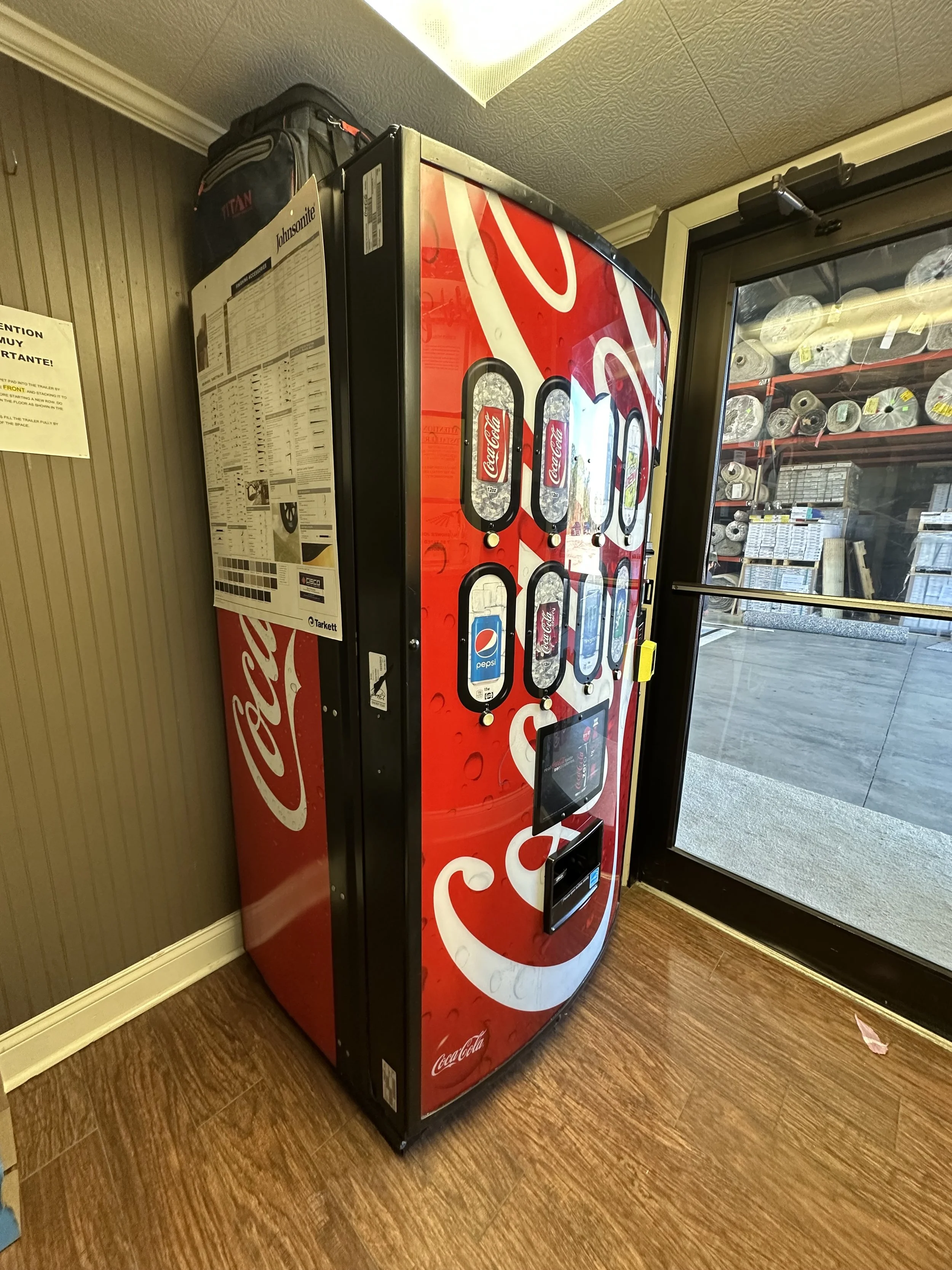 Coca-Cola vending machine next to a glass door in a convenience store, with a display screen and product selection buttons.