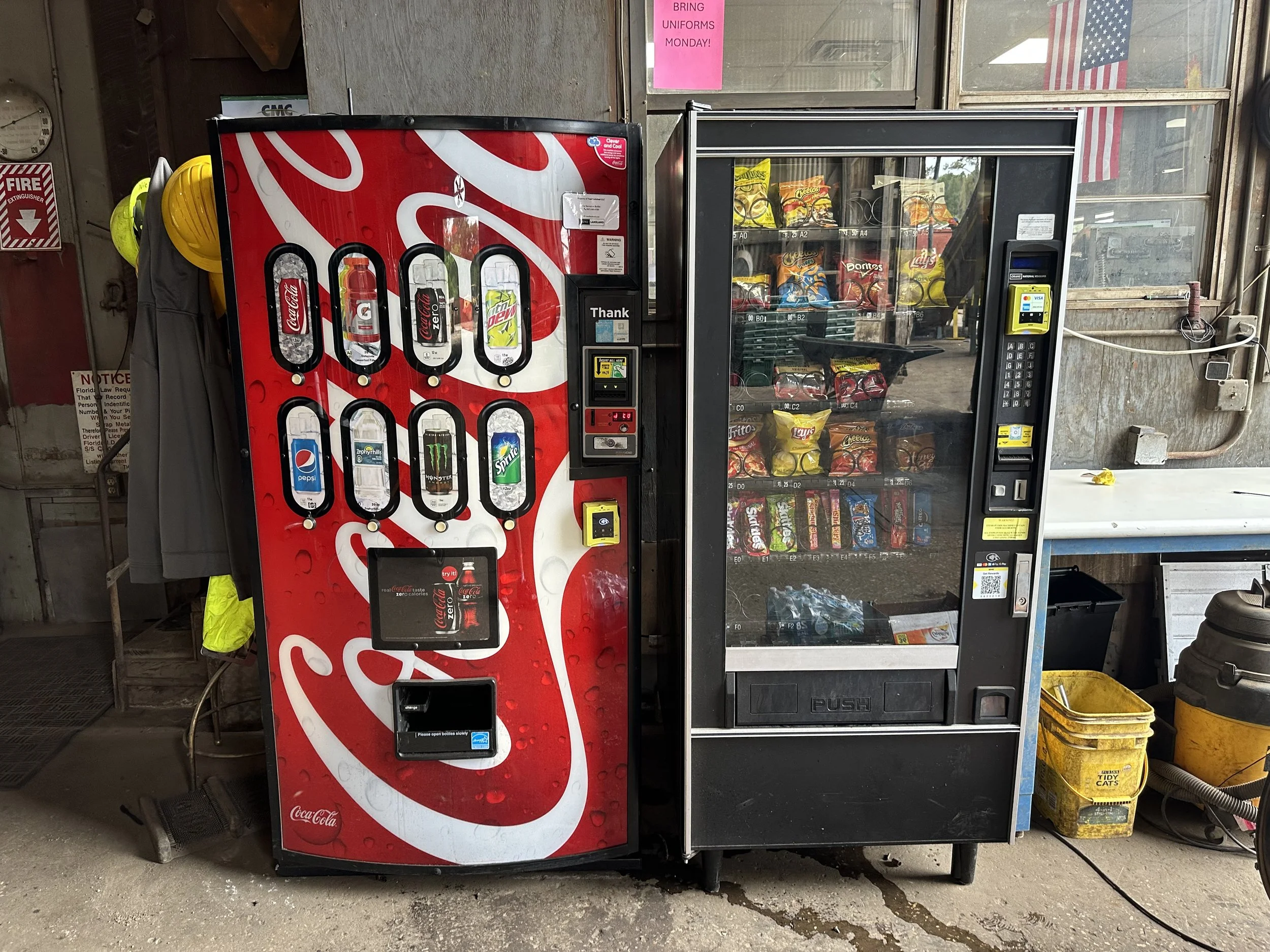 A Coca-Cola vending machine and a snack vending machine side by side in a store. The Coca-Cola machine has a red design with beverage options and a screen, while the snack machine displays chips and bottled water behind a glass door.
