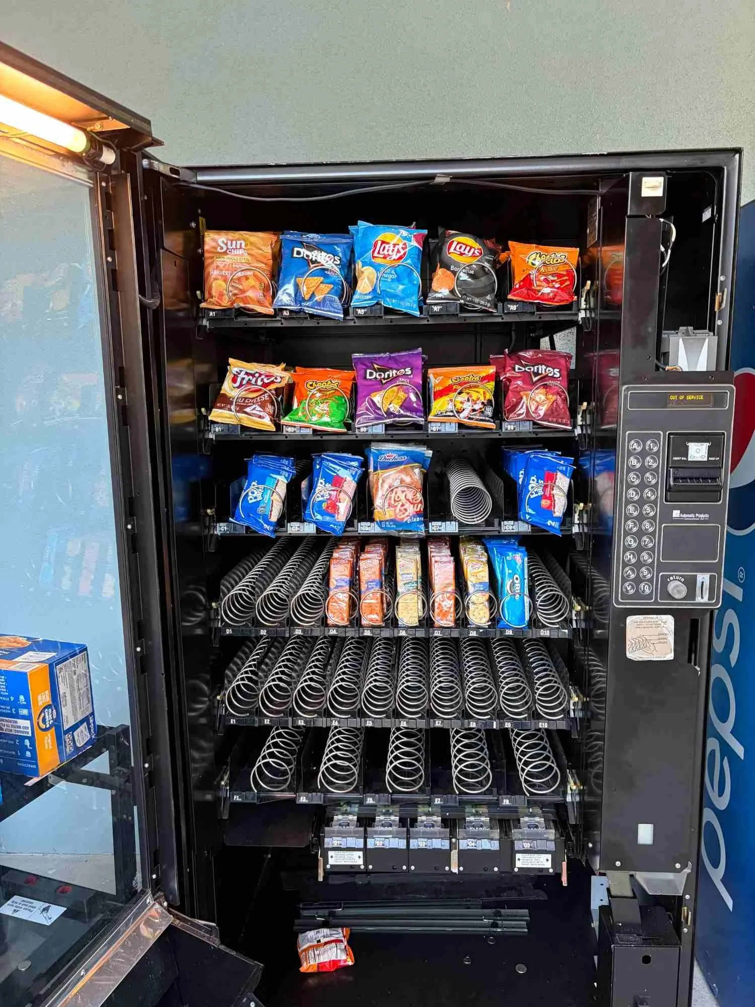 A vending machine with snacks, mostly chips, and no products inside, with some snack packages on the shelves and a snack bag on the floor.