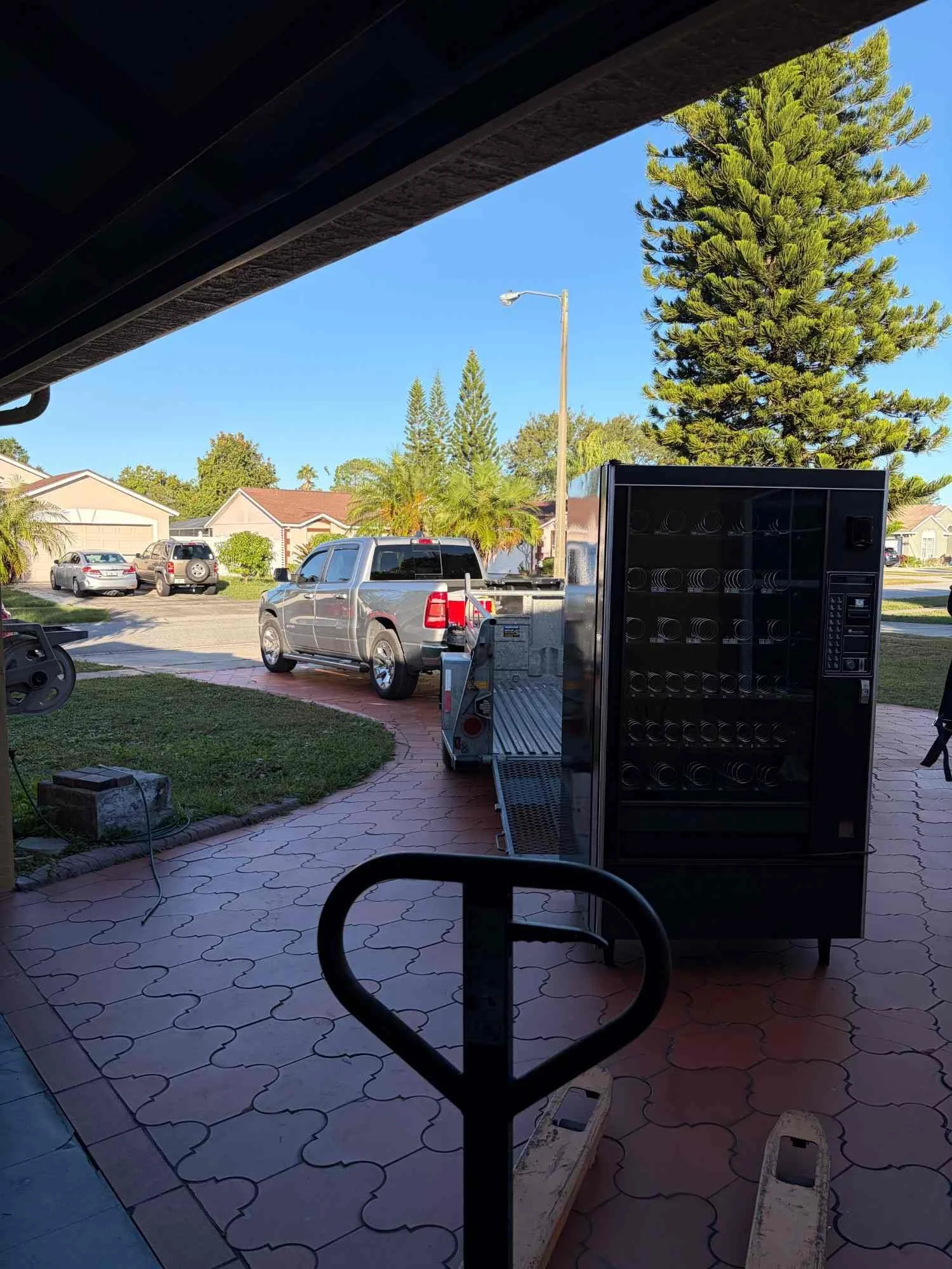 A view from inside a garage or outdoor workspace looking out onto a driveway with parked cars, a streetlamp, and a large pine tree, during daytime with clear blue sky.