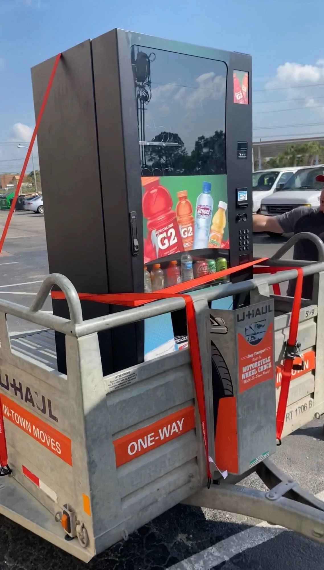 A vending machine inside a shopping cart in a parking lot, with a blue sky and trees in the background. The vending machine displays various beverages, including G2 low-calorie drinks, water, and soda.