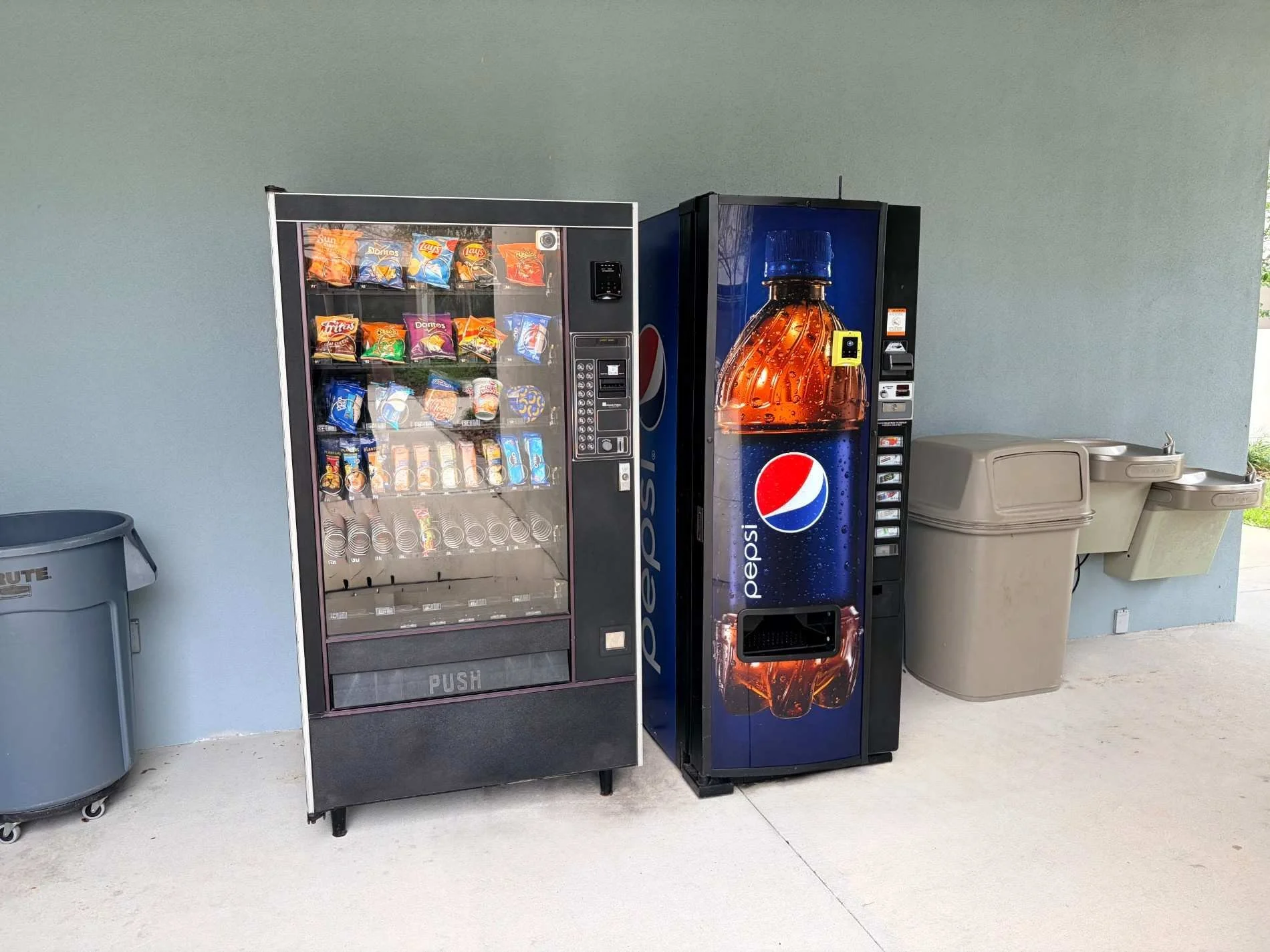 Two vending machines placed side by side against a light-colored wall. The left vending machine contains snack bags, and the right vending machine displays a large Pepsi soda bottle on its front.