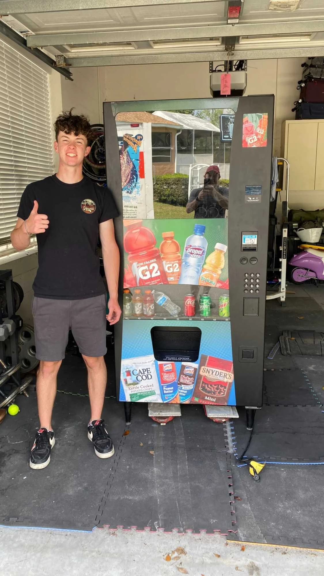 A young man with curly hair giving a thumbs-up sign, standing next to a vending machine outside in a garage or storage area.