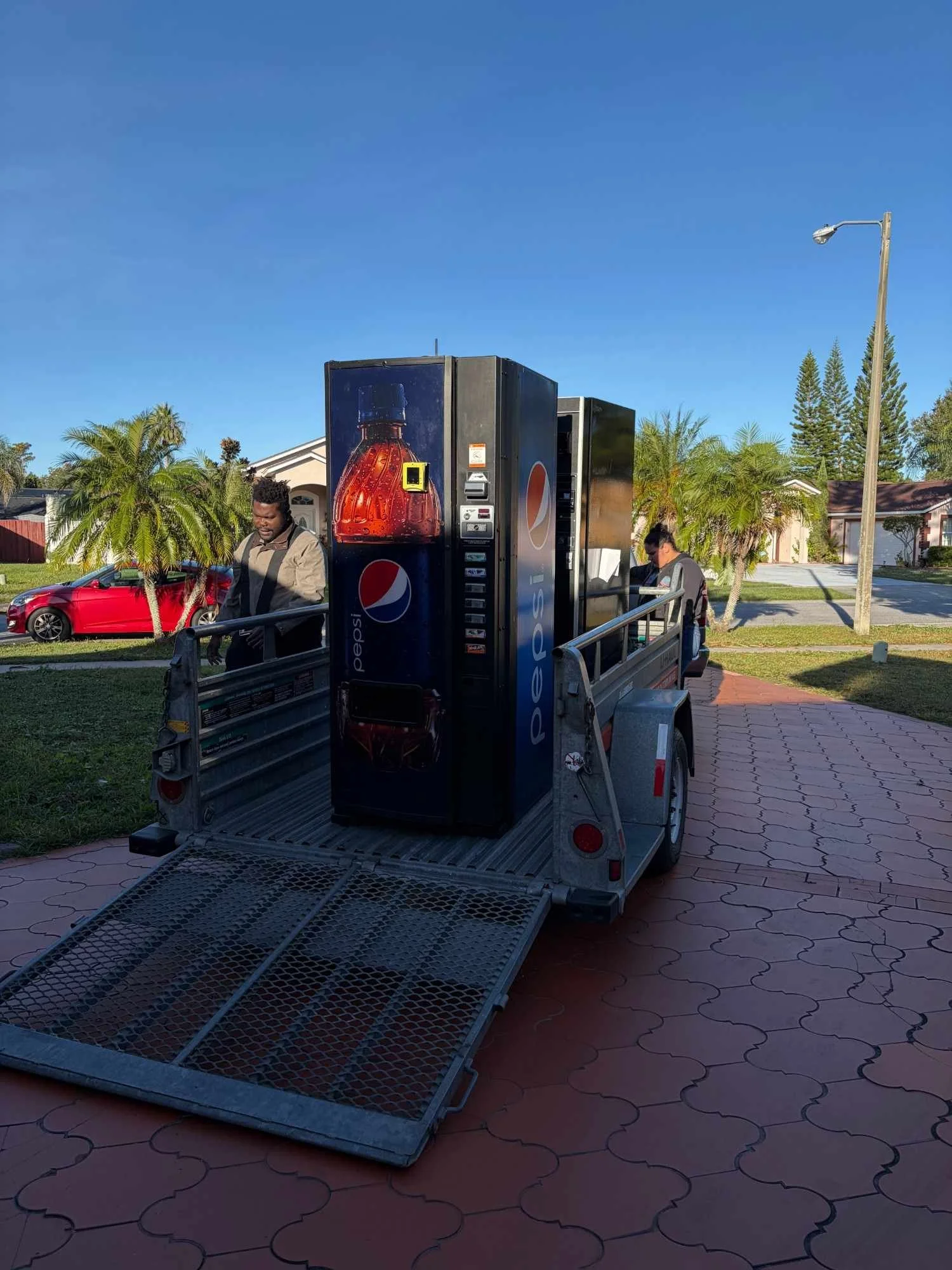 A small truck with a Pepsi vending machine on its bed parked on a sidewalk in a suburban neighborhood. Two people are near the vending machine, one man and one woman, with houses and trees in the background and a bright blue sky above.