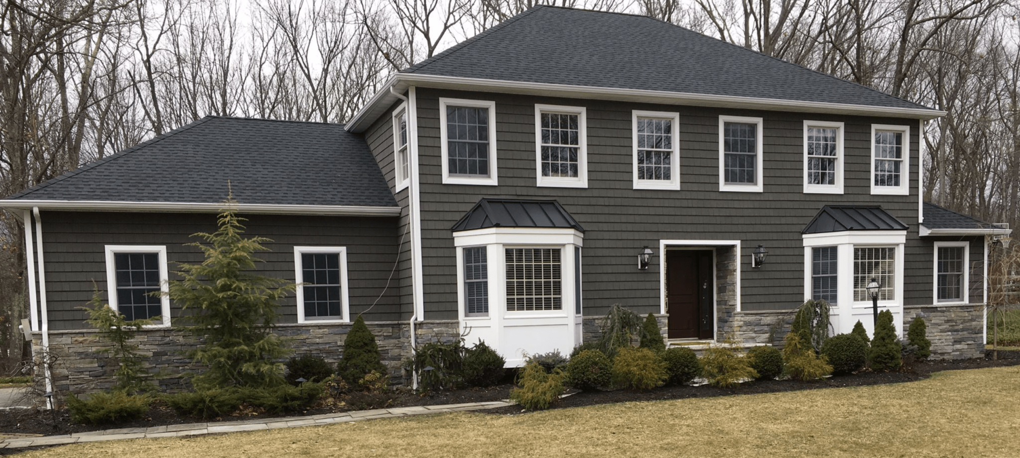 Front view of a two-story house with dark gray siding, white window frames, black roof, stone accents at the base, and landscaped bushes and trees in front.