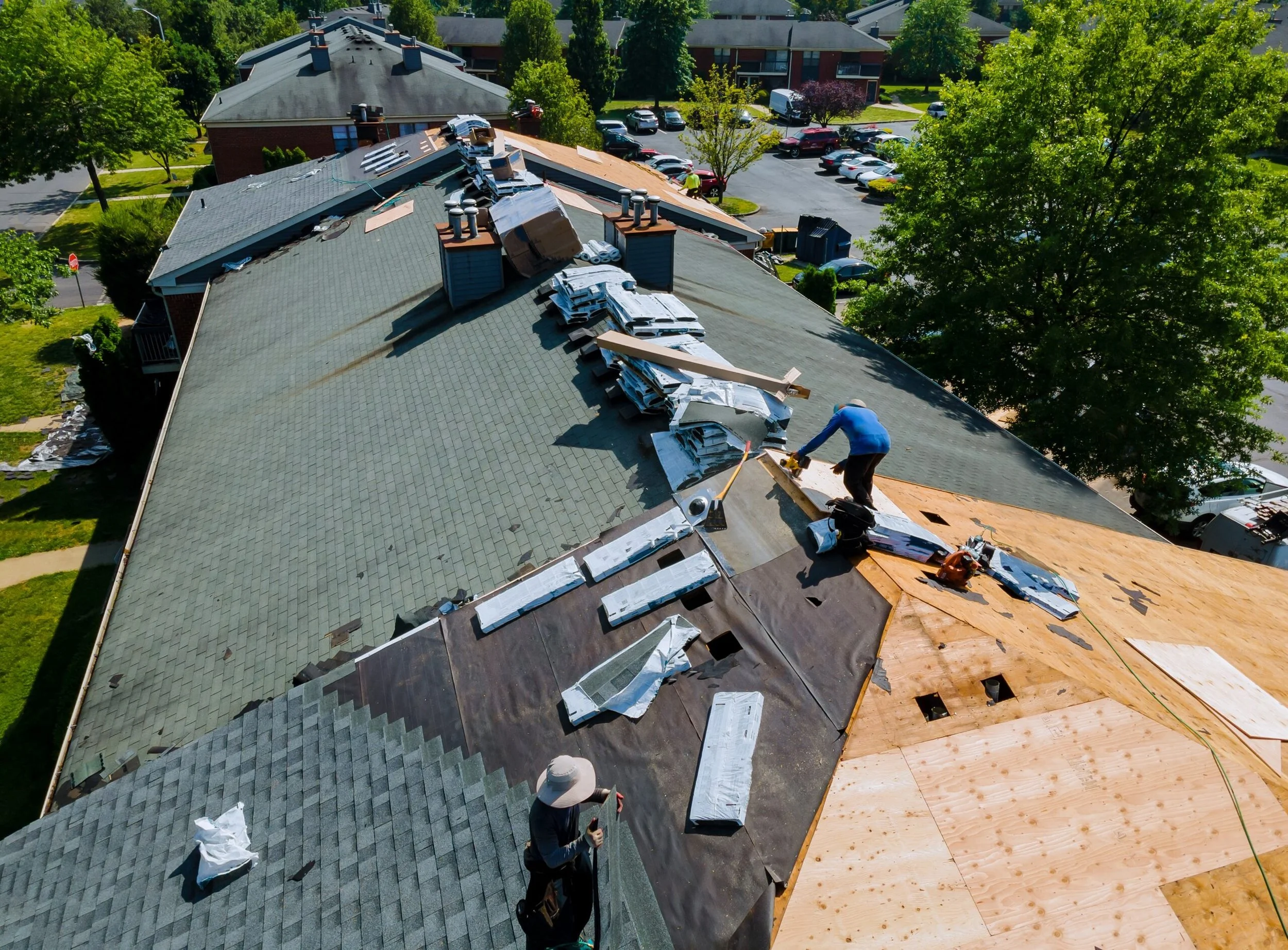 Workers installing new roofing on a large building, with shingles and plywood, in a suburban neighborhood with trees and parking lot visible.