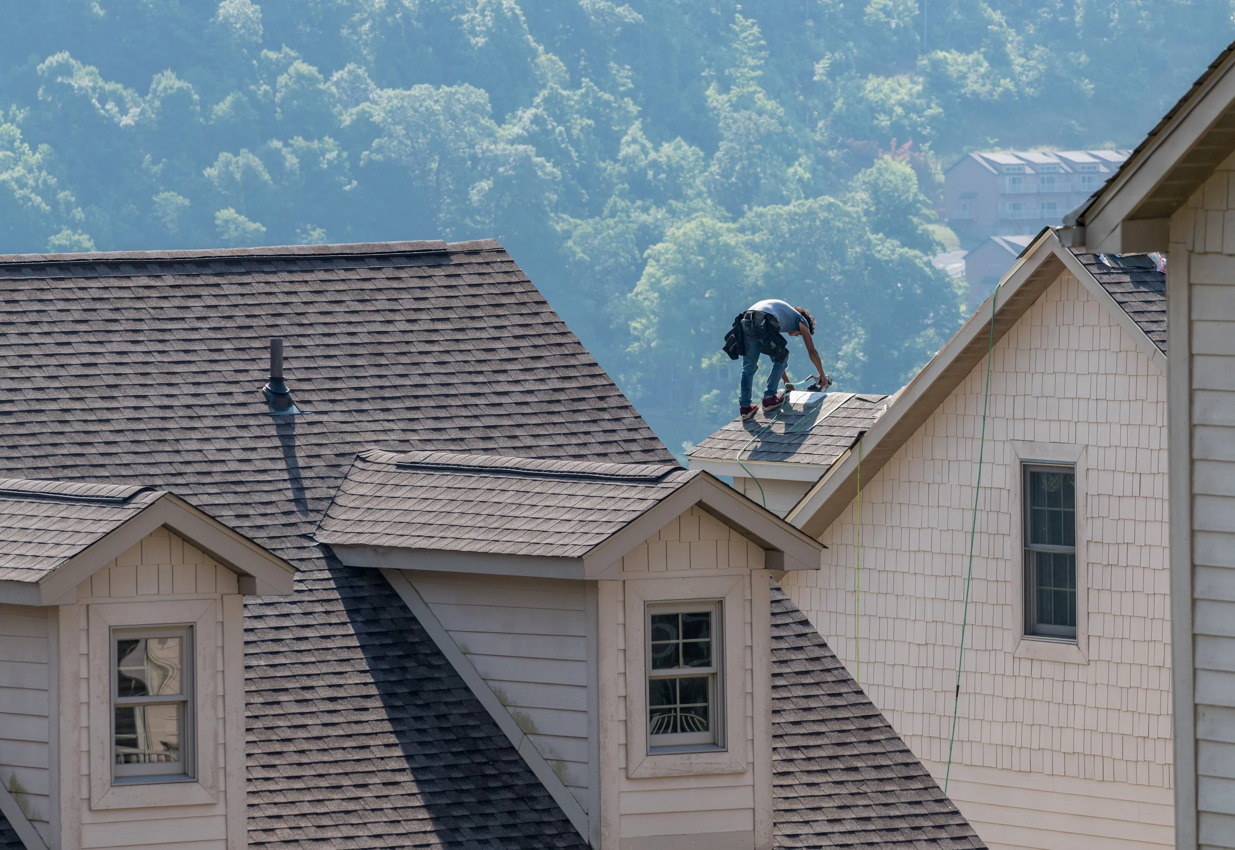 A person working on the roof of a house, repairing or inspecting shingles on a sunny day with green trees and other houses in the background.