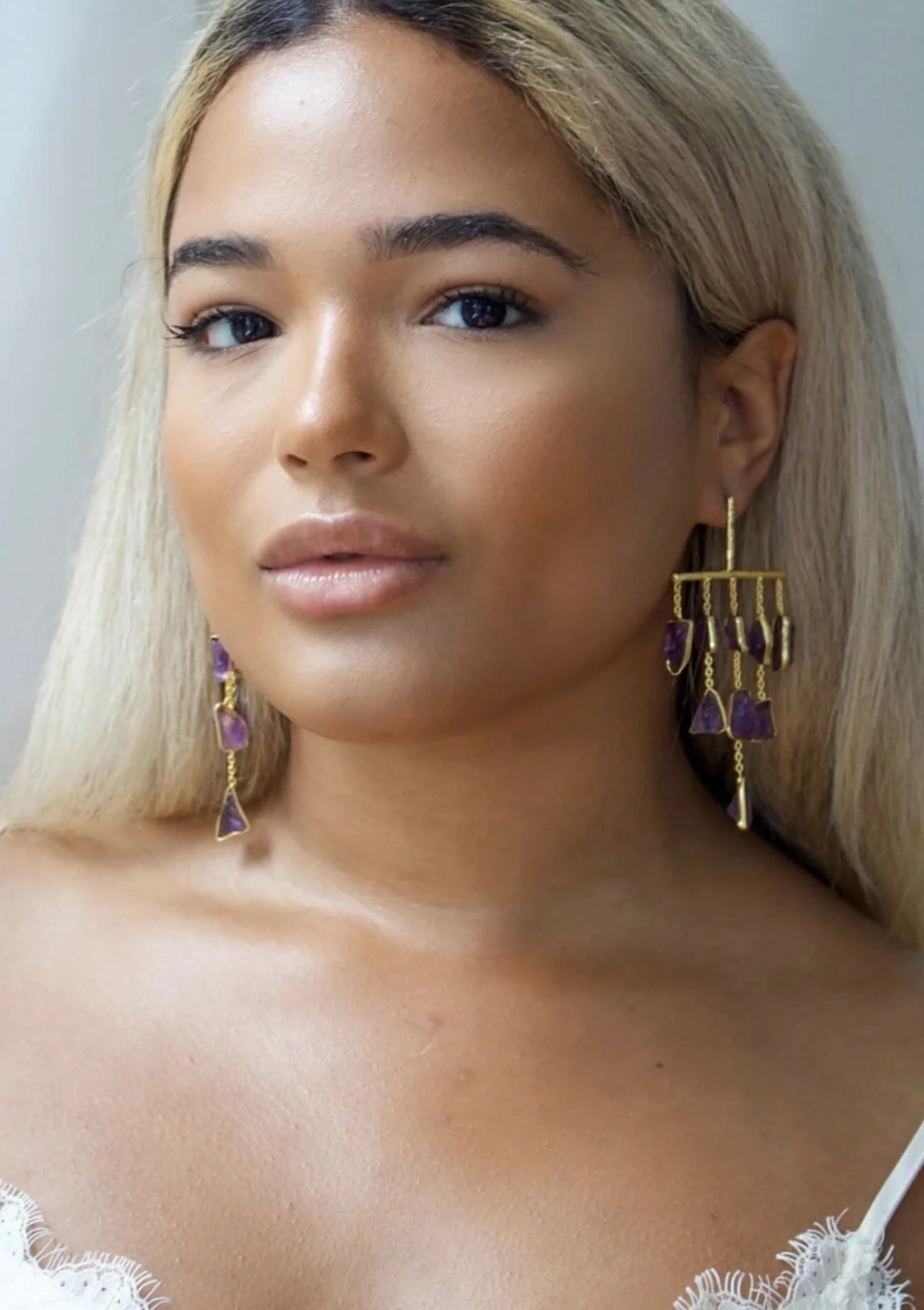 Close-up of a woman with long blonde hair, wearing large gold earrings with purple stones, and a white lace top.