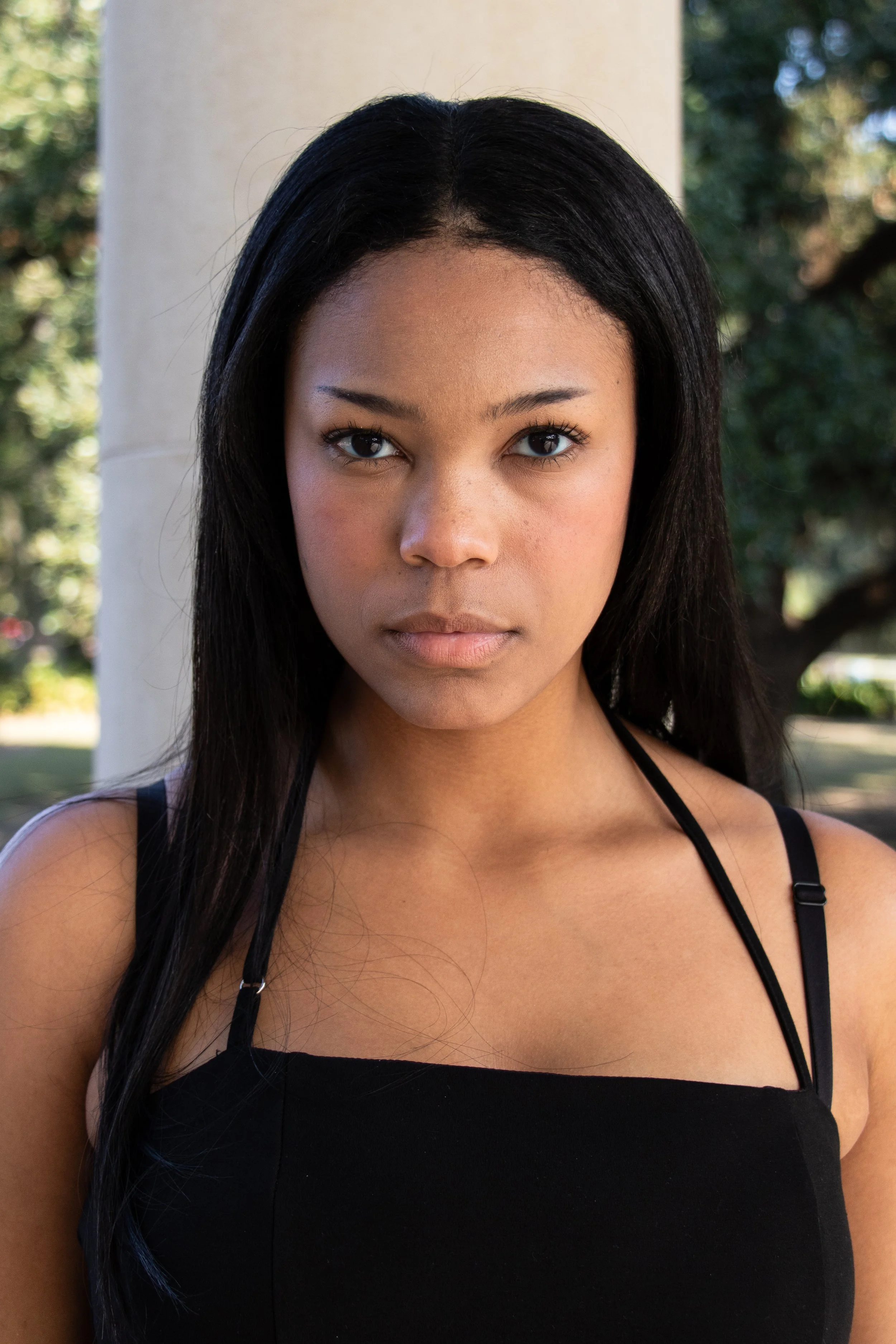 Close-up of a young woman's face with long black hair, wearing a black top with thin straps, standing outdoors in front of a white column and blurry trees in the background.