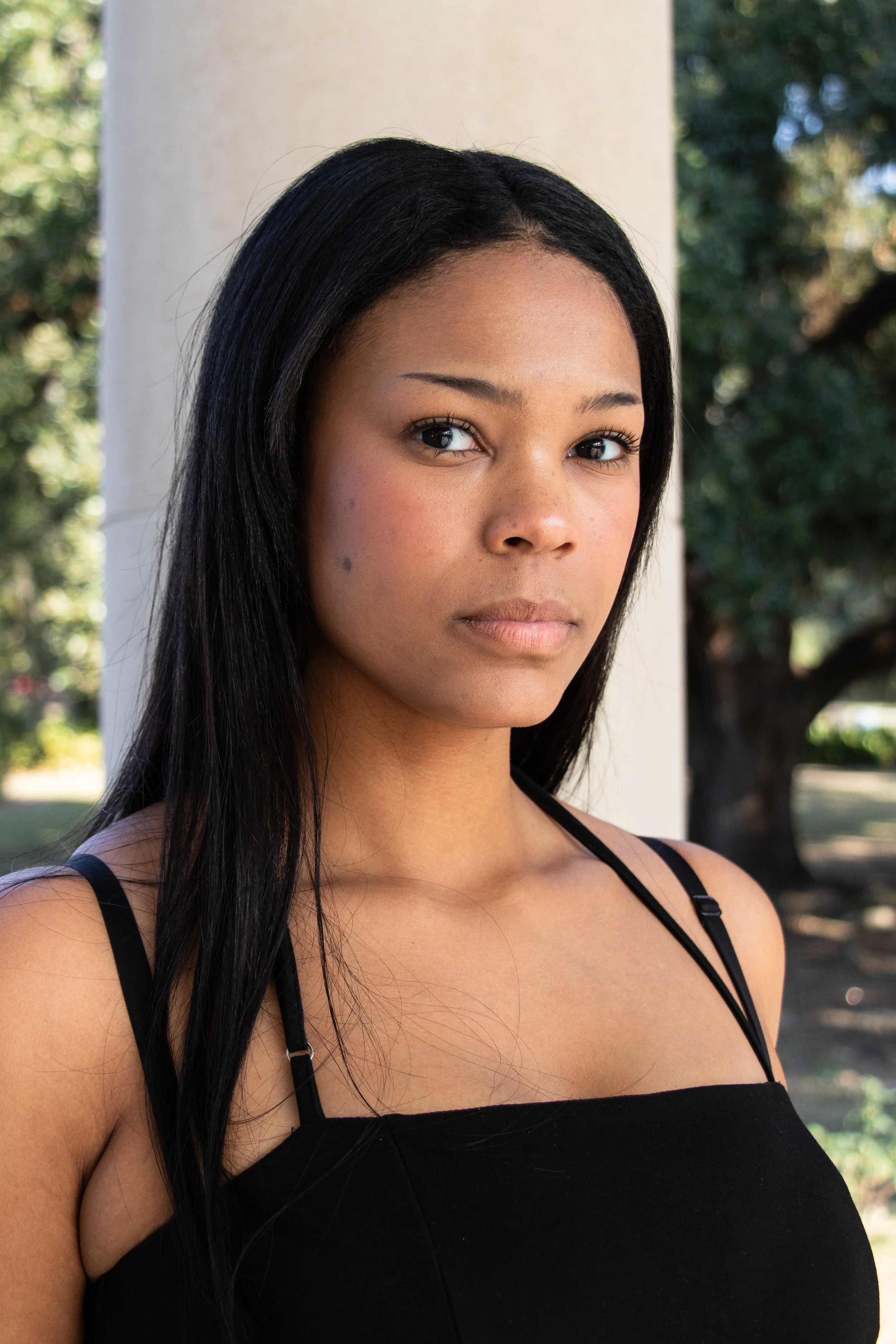 A young woman with long black hair and tan skin standing outdoors near a white column, with trees and greenery in the background, wearing a black spaghetti strap top.