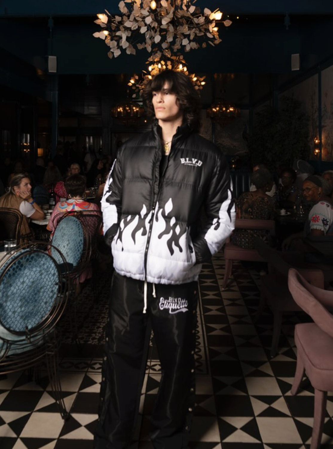 A young man with dark, curly hair wearing a black and white puffer jacket and matching black pants standing in a dimly lit restaurant with chandeliers, patterned floor, and seated patrons in the background.