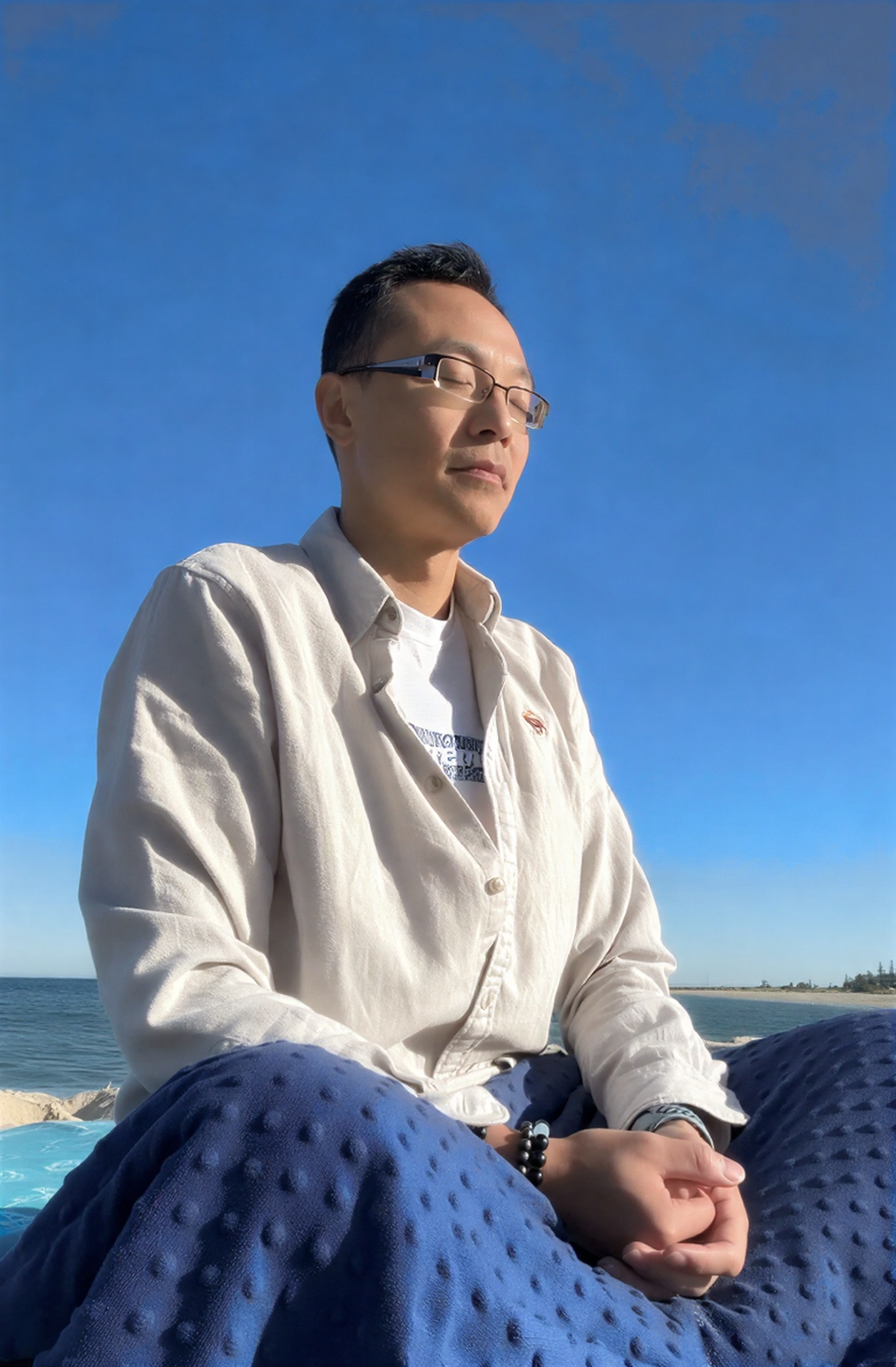 A man with short dark hair and glasses meditating near a beach, eyes closed, wearing a light-colored shirt, sitting on a blue textured blanket with the ocean and a distant shoreline in the background.