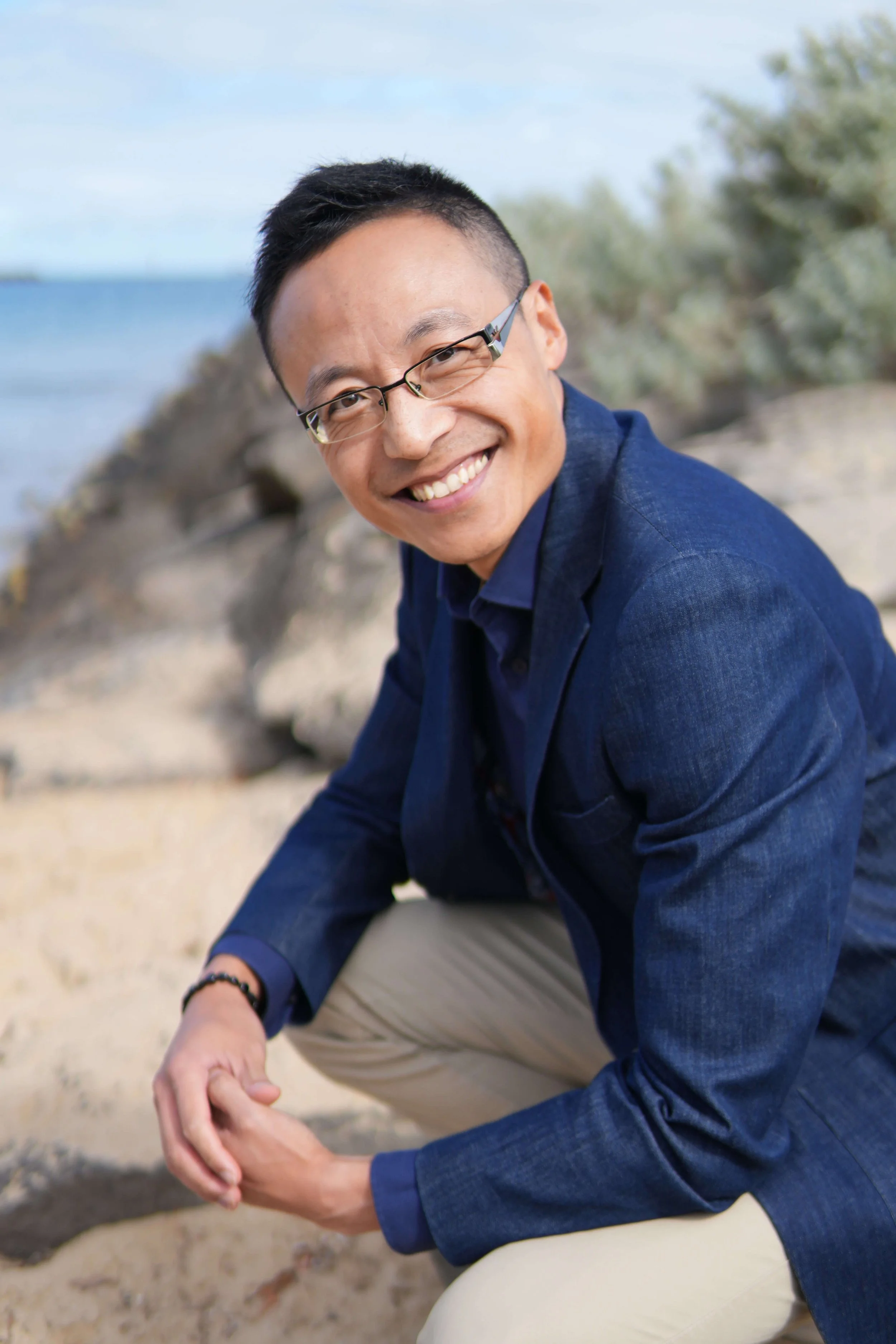A man with glasses and short dark hair smiling at the camera while sitting on a beach, wearing a blue jacket and beige pants.