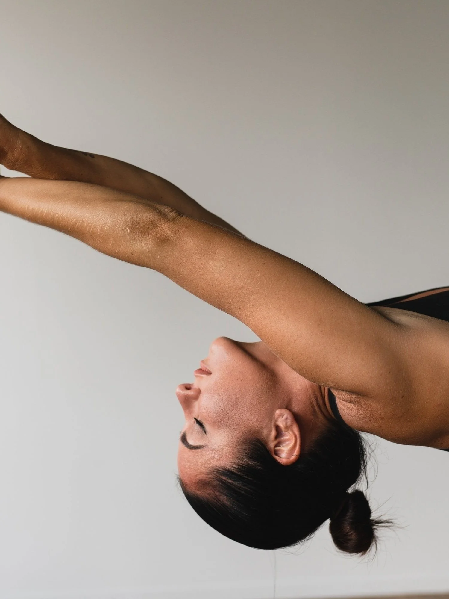 A woman with dark hair tied in a bun stretching with her arms extended overhead, side profile visible, against a plain neutral background.