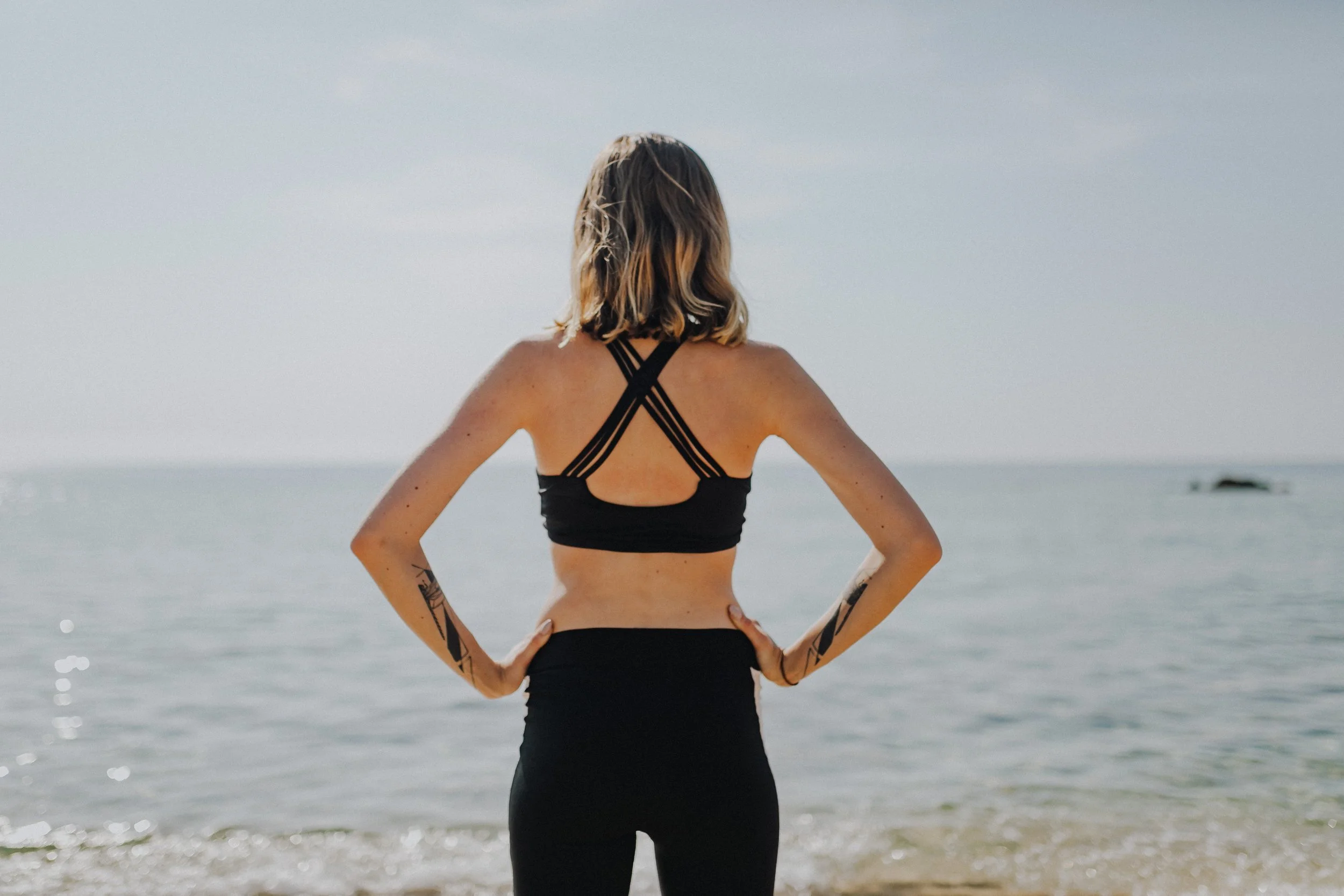 A woman with short blonde hair standing on a beach, facing the water, wearing a black sports bra and black pants, with minimal tattoos on her arms.