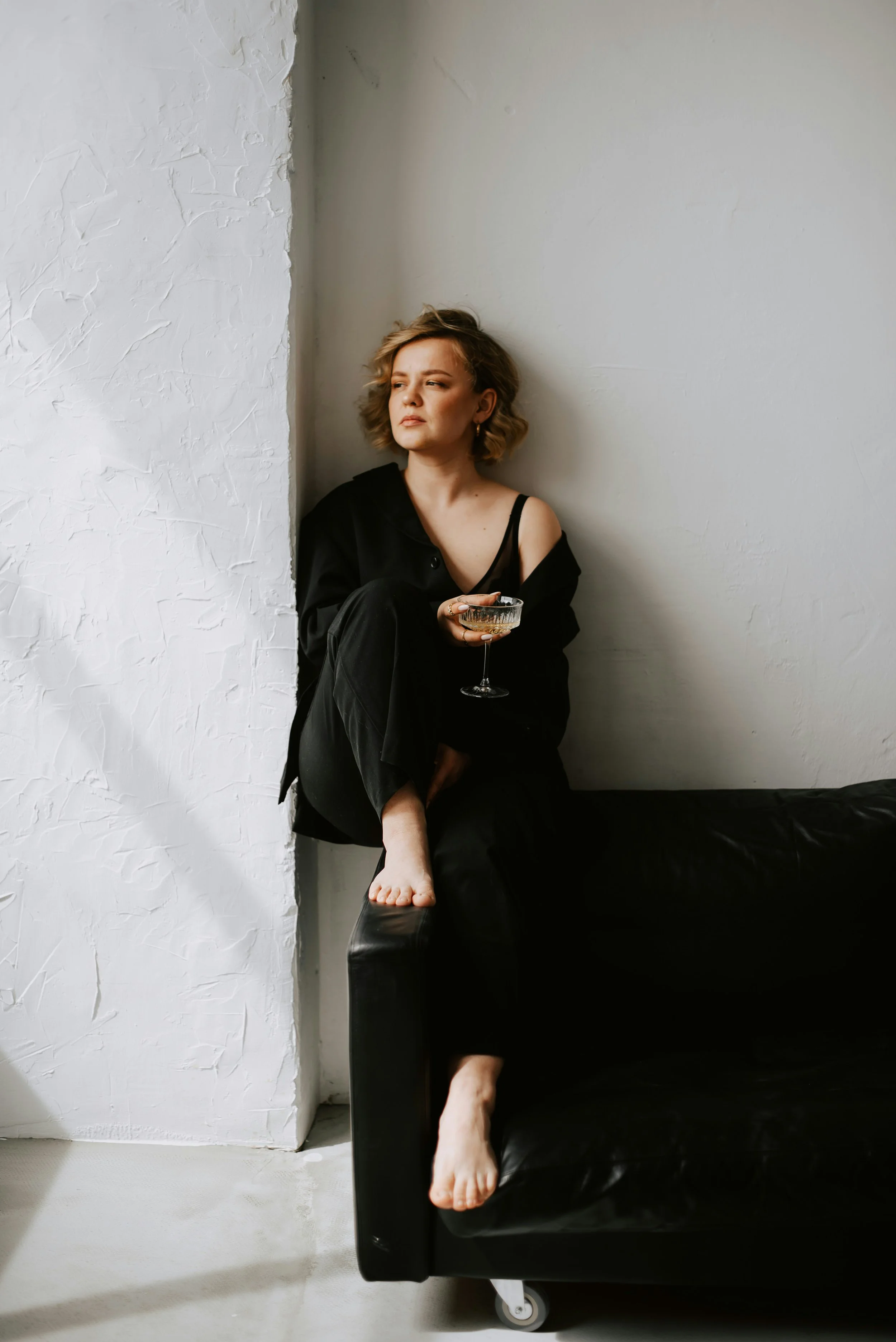 A woman with short wavy hair, wearing a black dress and a black jacket, sitting on the armrest of a black sofa in a minimalistic room with white textured walls, holding a glass of champagne, looking contemplative.