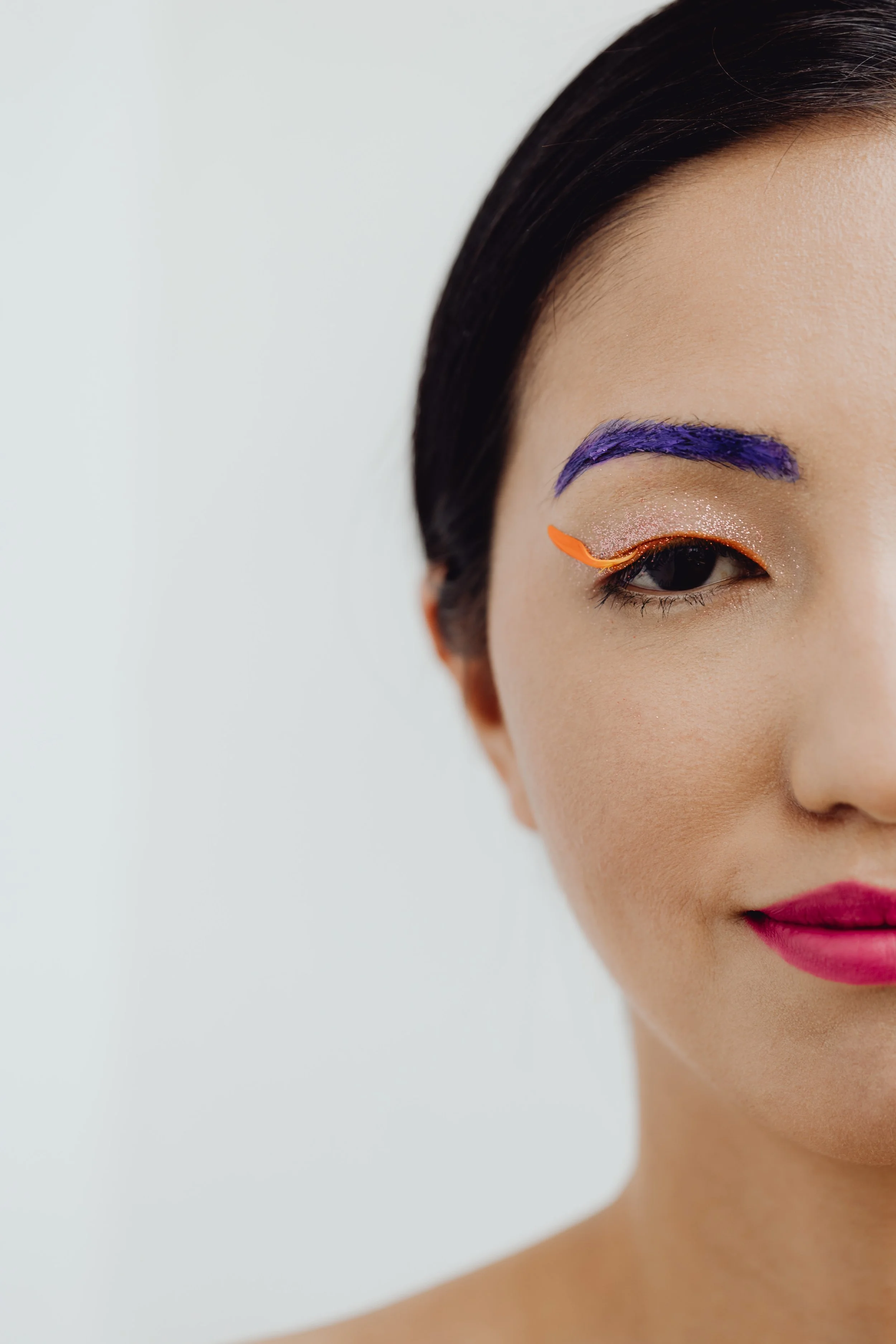 Close-up of a woman with makeup styling, featuring bold purple eyebrows, shimmery eyeshadow, bright orange eyeliner with a feather-like tip, and vibrant pink lipstick, against a plain white background.