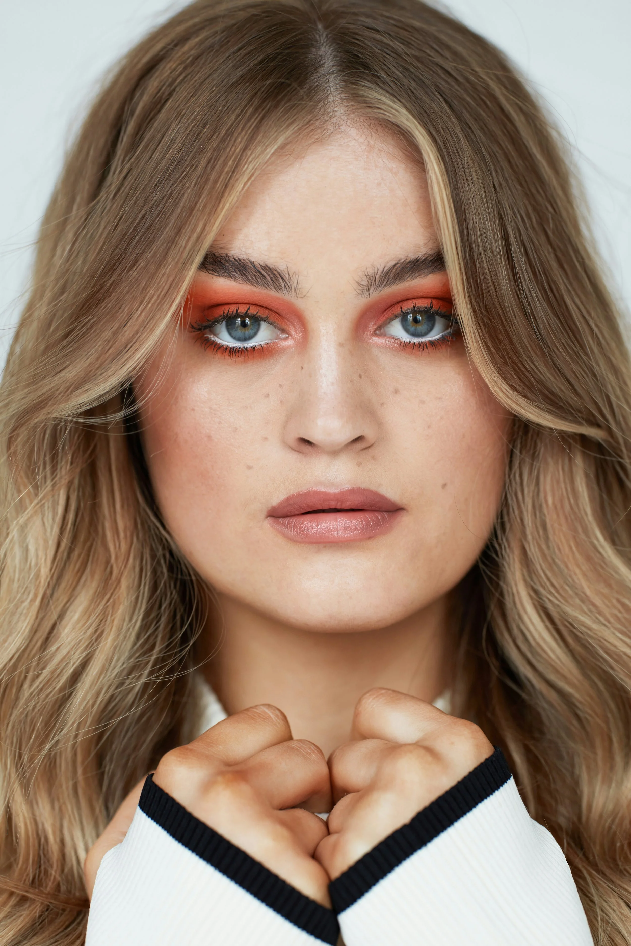 Close-up portrait of a young woman with long wavy blonde hair, blue eyes, wearing orange eye makeup, and a white top with black accents, holding her hands together near her chest.