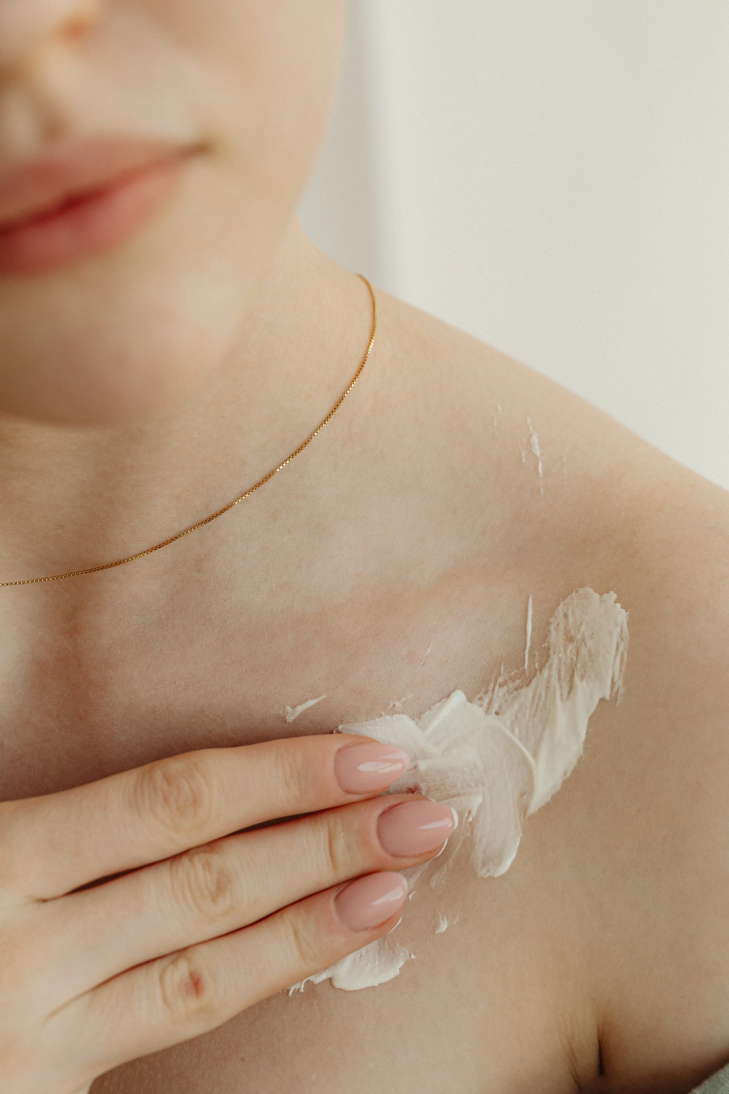 Person applying lotion to their shoulder with a hand with manicured nails, wearing a delicate gold necklace.