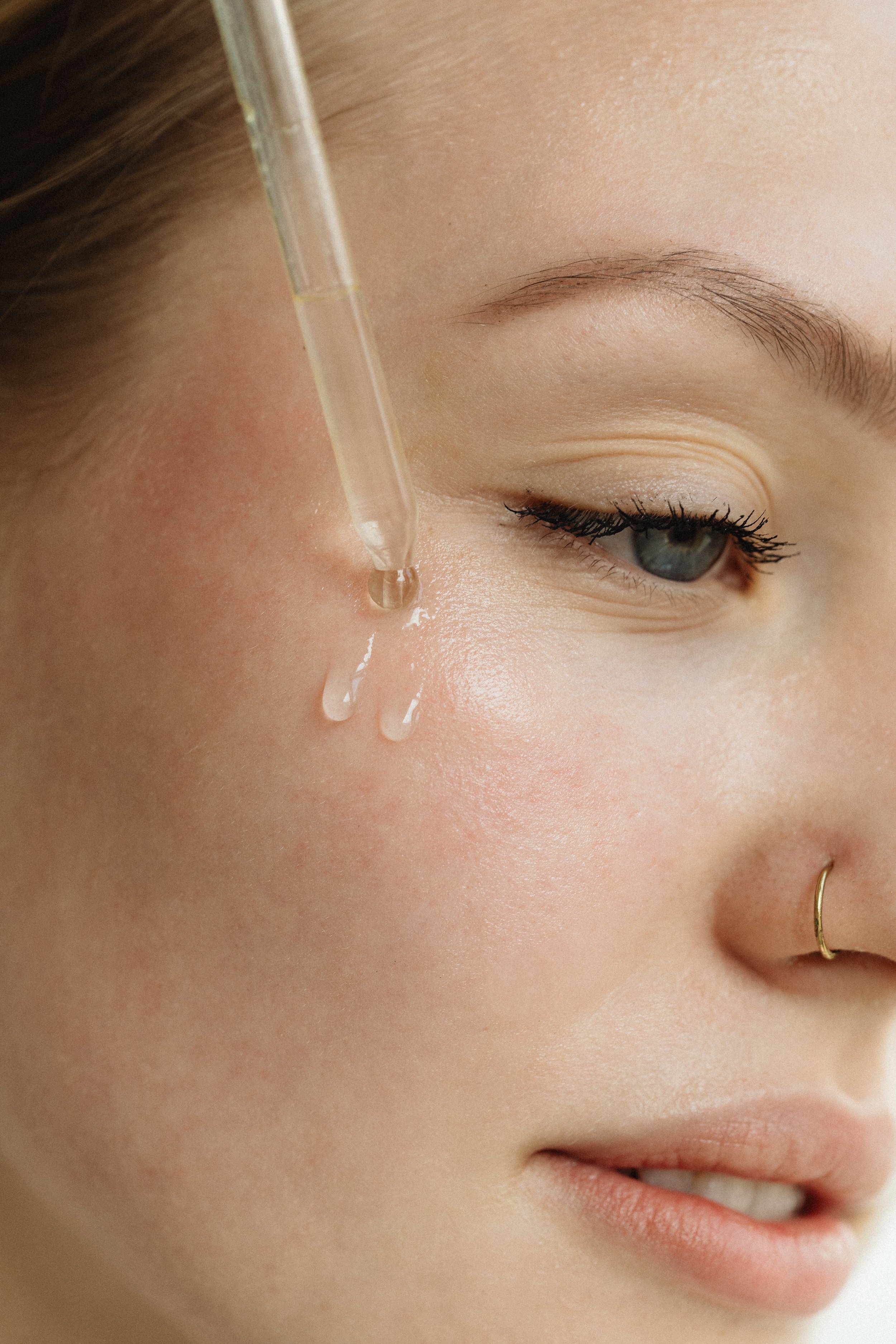 Close-up of a woman with a nose ring applying serum to her face with a dropper.