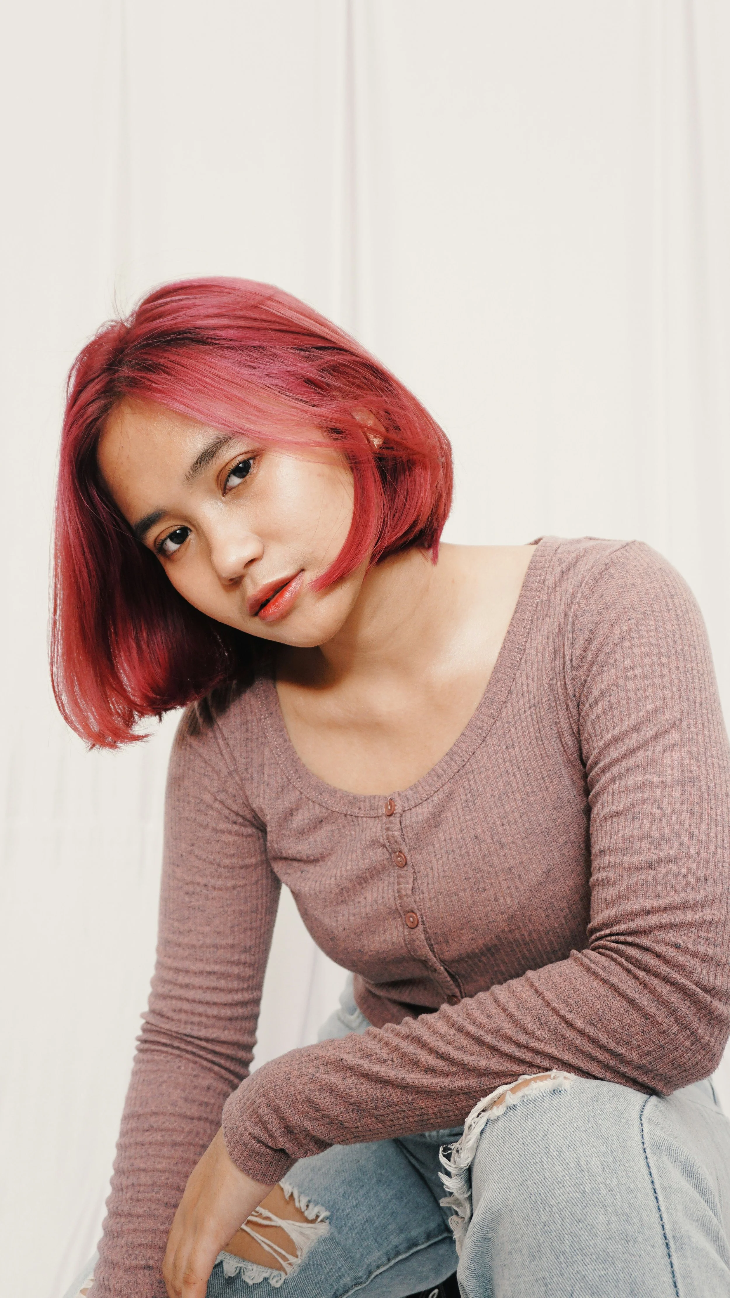 A young woman with straight, shoulder-length pink hair, wearing a long-sleeve brown shirt with buttons and ripped jeans, sitting against a plain light-colored wall, looking at the camera.