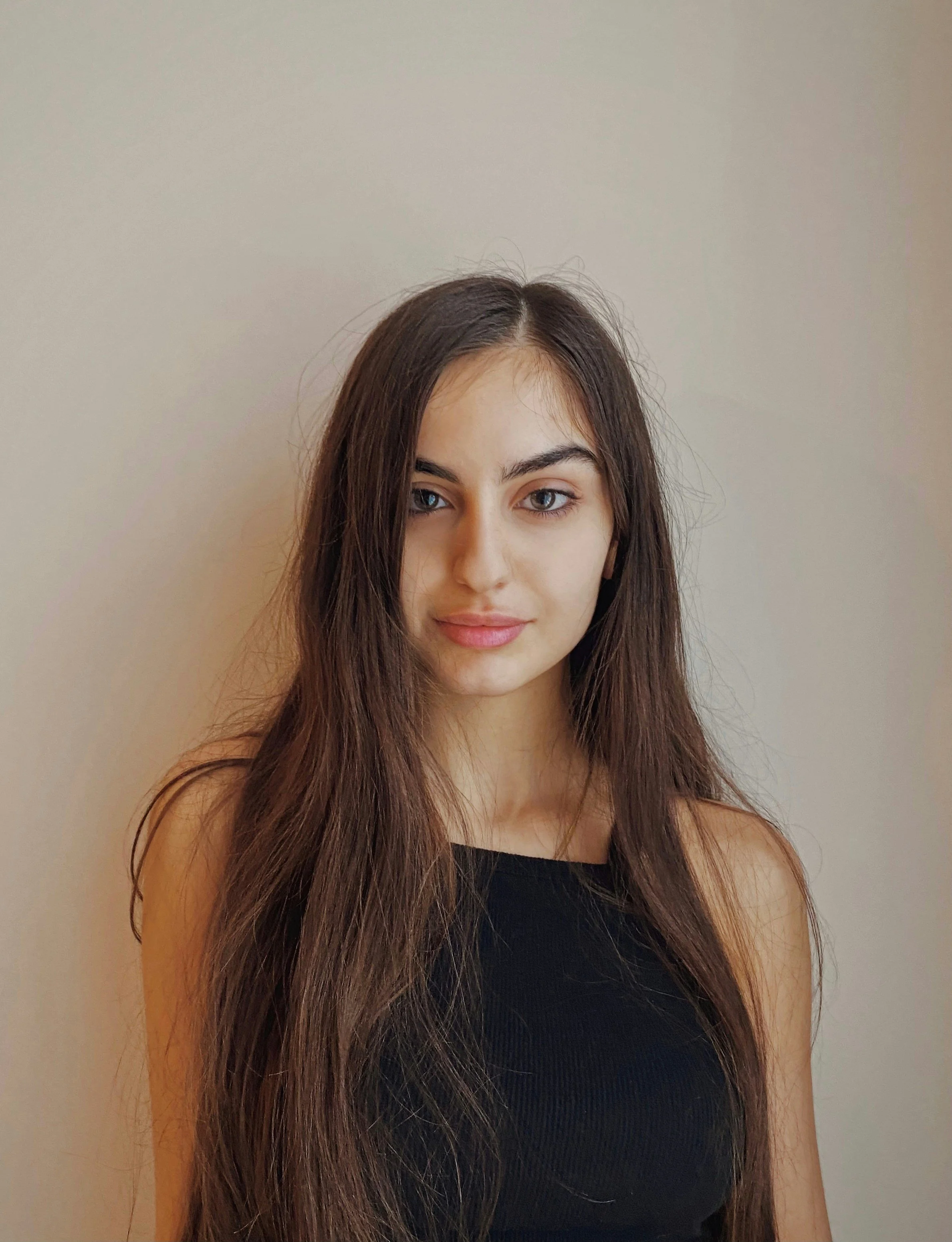 Young woman with long brown hair and blue eyes, wearing a black sleeveless top, standing against a plain wall