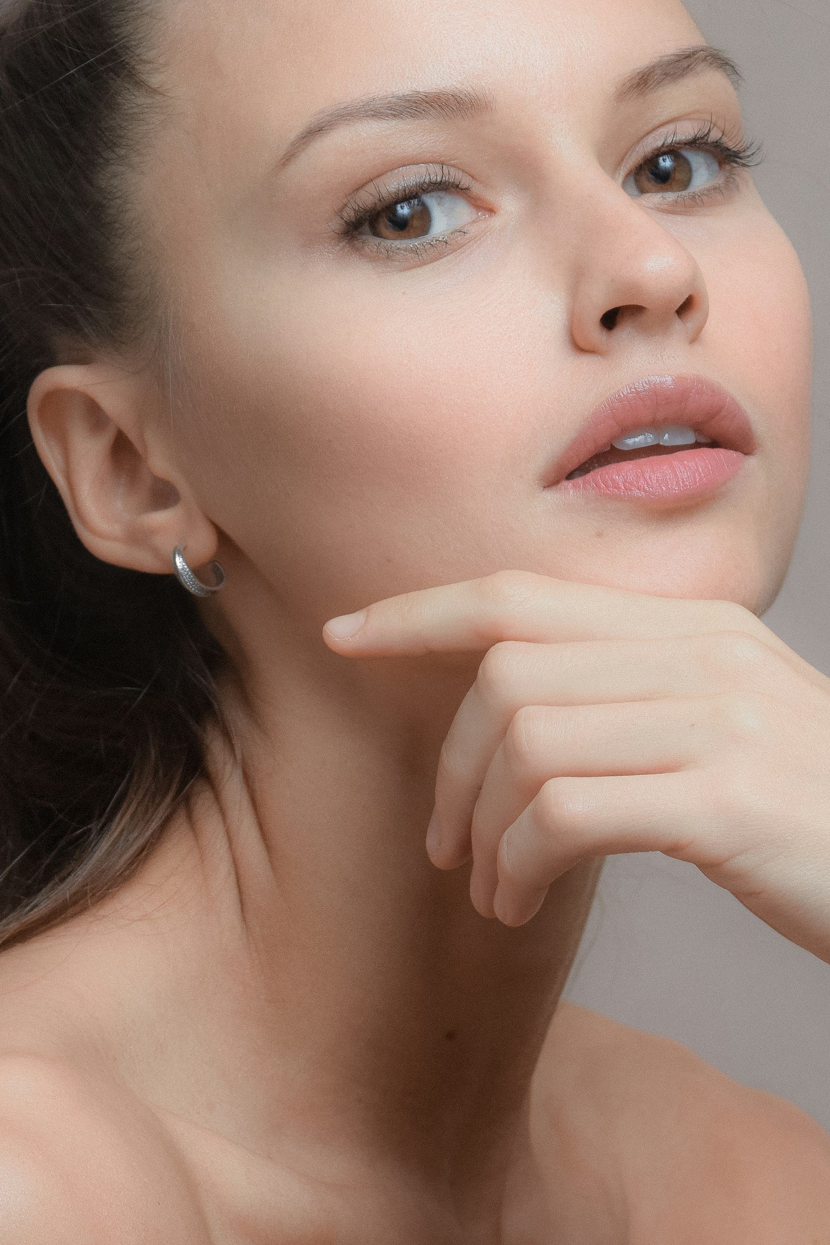 Close-up portrait of a young woman with flawless skin, light makeup, blue eyes, and brown hair, touching her chin gently with her hand.