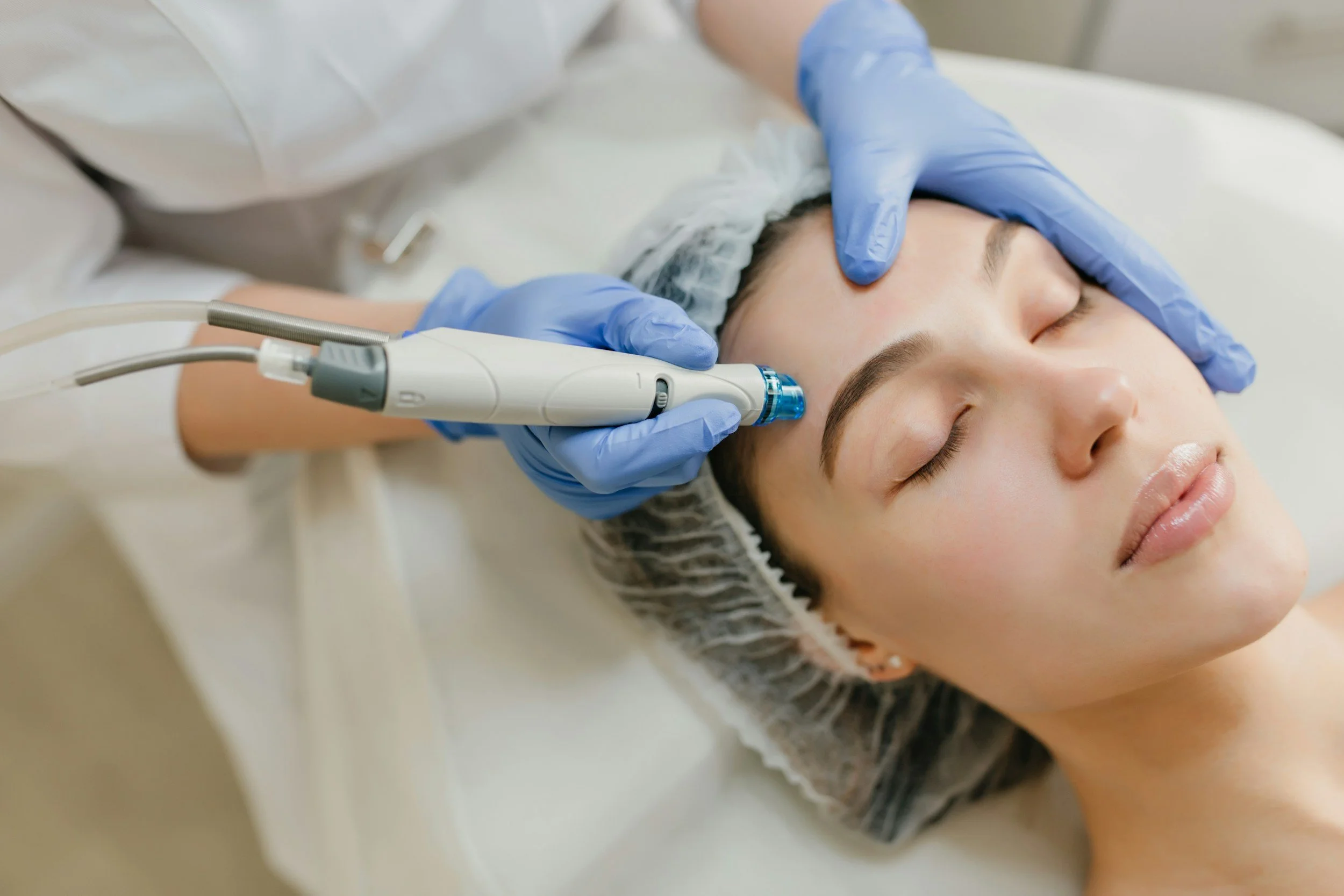 A woman receiving a facial treatment with a handheld device, lying down with eyes closed, wearing a hair cap, in a clinical setting.