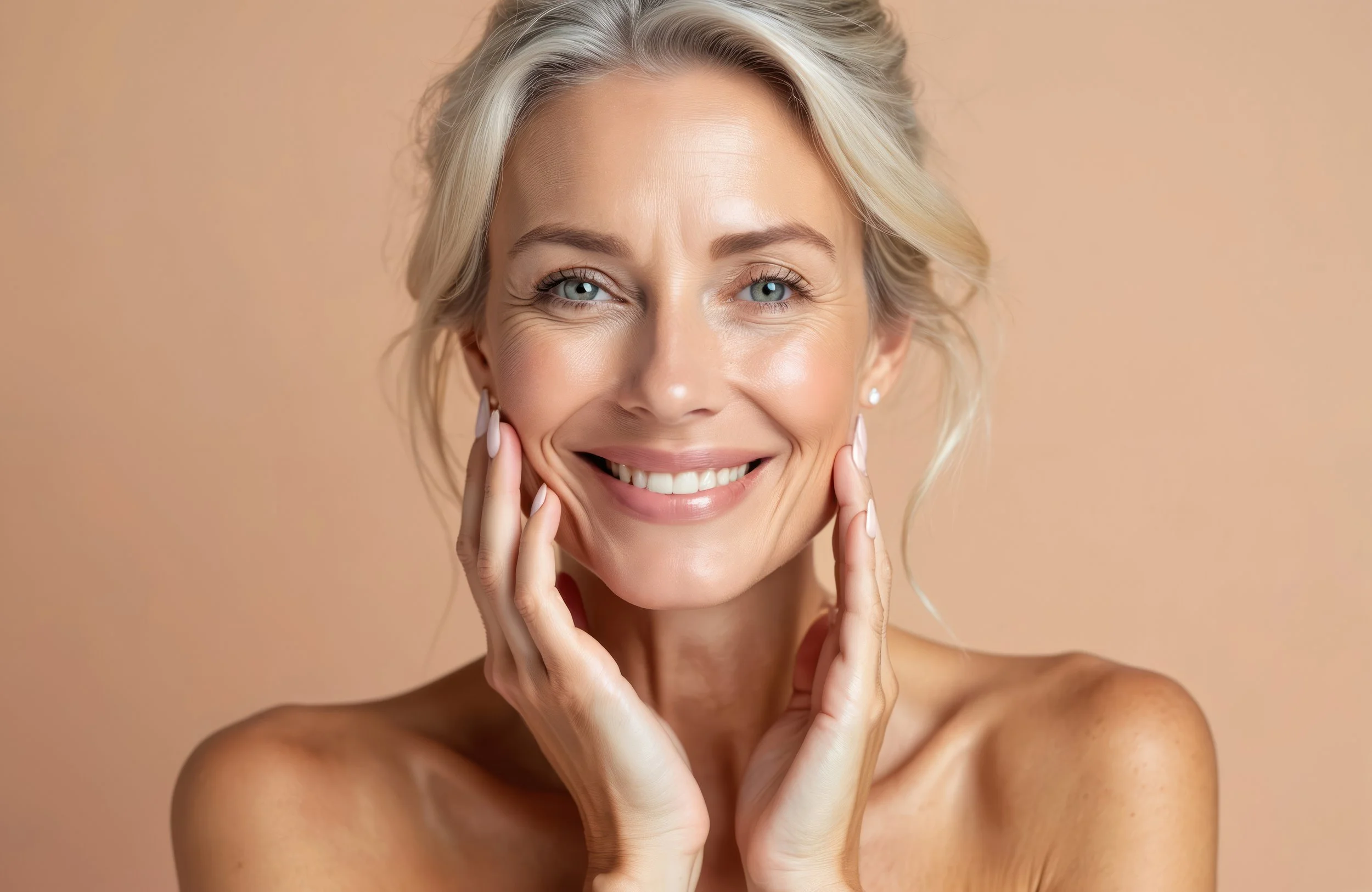 A smiling woman with blonde hair and blue eyes, touching her face gently, with bare shoulders, against a soft beige background.