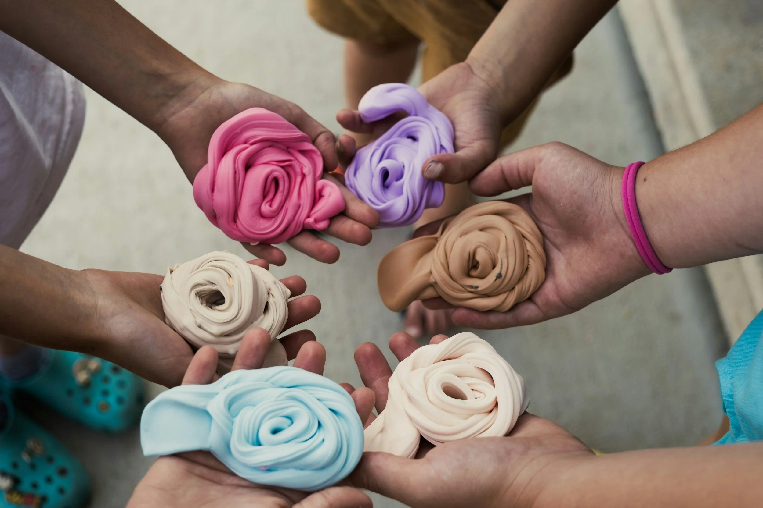 Multiple hands hold colorful spiral-shaped objects that resemble rolled fabrics or soft sculptures, in shades of pink, purple, beige, white, and blue.