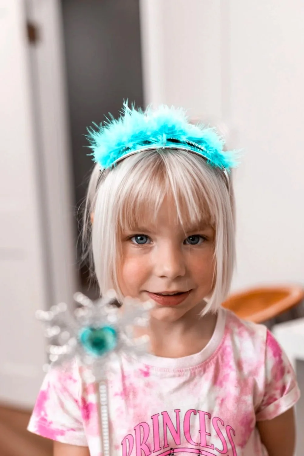 A young girl with blonde hair and blue eyes wearing a pink tie-dye shirt that says 'Princess' and a fluffy blue headband. She is smiling and holding a sparkly wand with a decorative top.