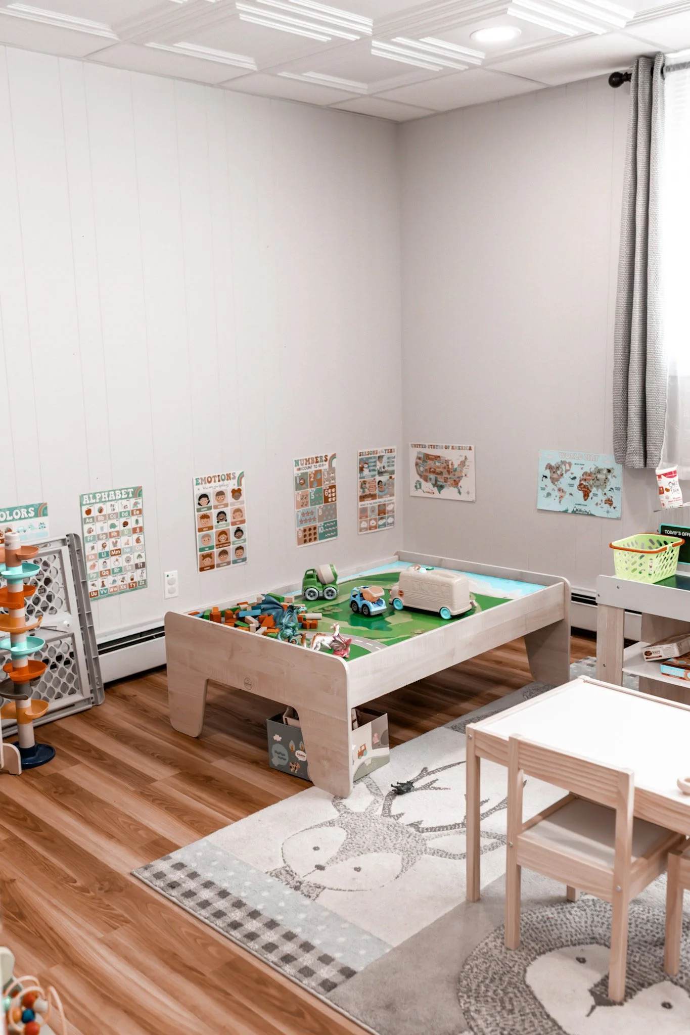 A children's playroom with a wooden bed and educational posters on the wall. There are toy cars, a small table with two chairs, and a colorful rug with an animal face design. The room has hardwood floors and a window with curtains.